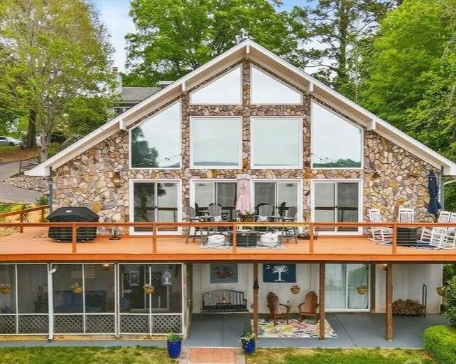 A large, multi-level house with a stone exterior, large glass windows, and a spacious wooden deck with outdoor furniture and a grill.