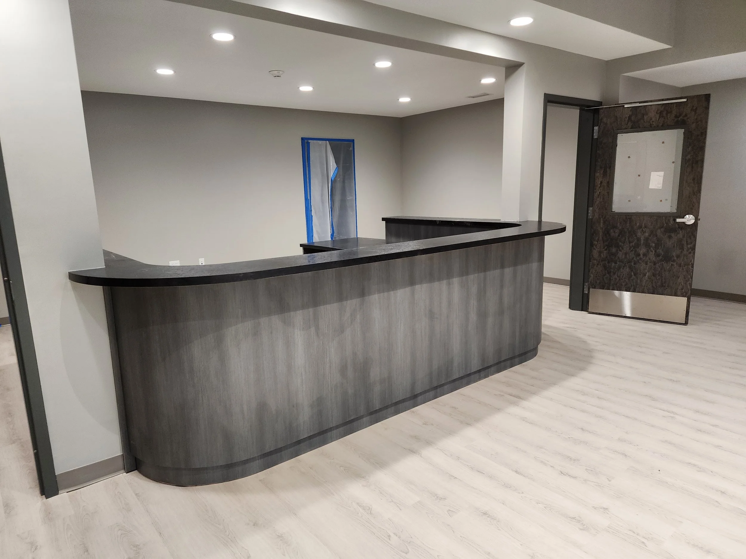 Modern reception desk with a dark countertop and wood-textured front panel in a well-lit room with white ceiling and light-colored wood flooring.