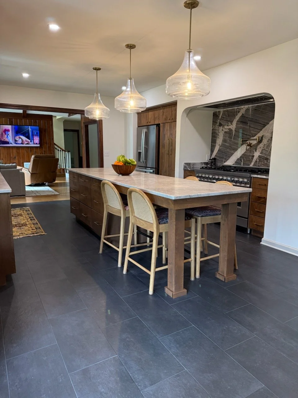 A modern kitchen with a central island, three pendant lights, a bowl of bananas and other fruit, dark tiled floor, wooden cabinets, stainless steel refrigerator, and a cooking area with a black marble backsplash.