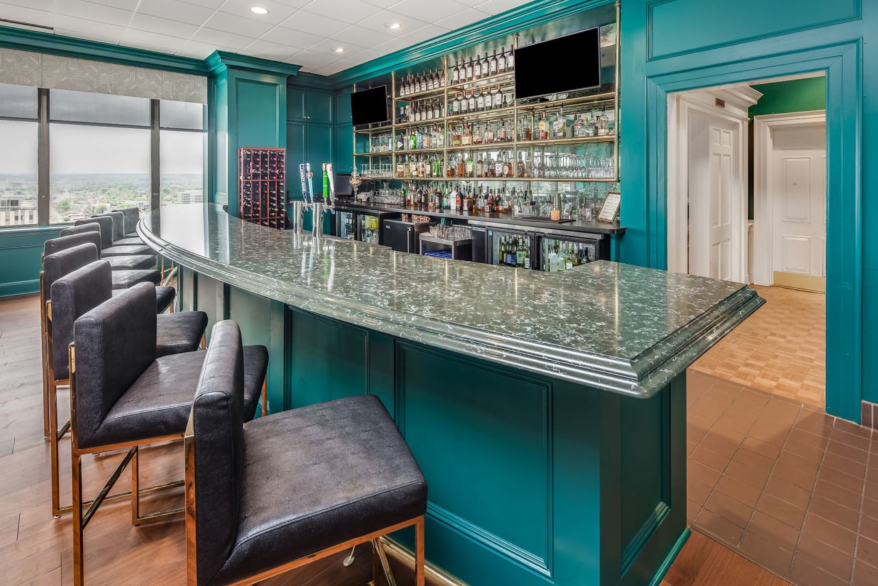 A stylish indoor bar with a green marble countertop, teal bar cabinets, and a wall lined with shelves of liquor bottles, with two television screens above.