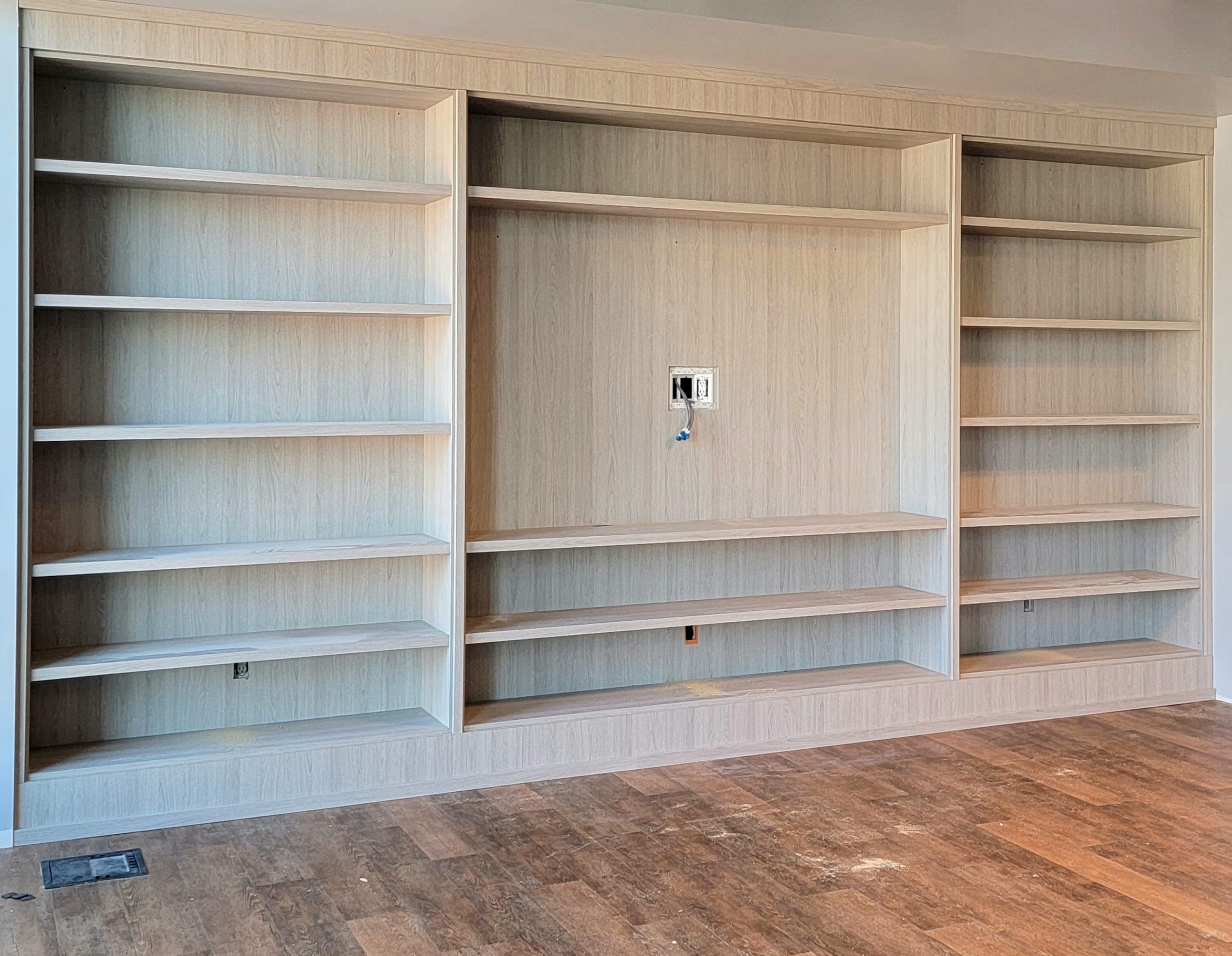 Empty wooden bookshelves with an electrical outlet and wiring on the wall, on a brown tiled floor.