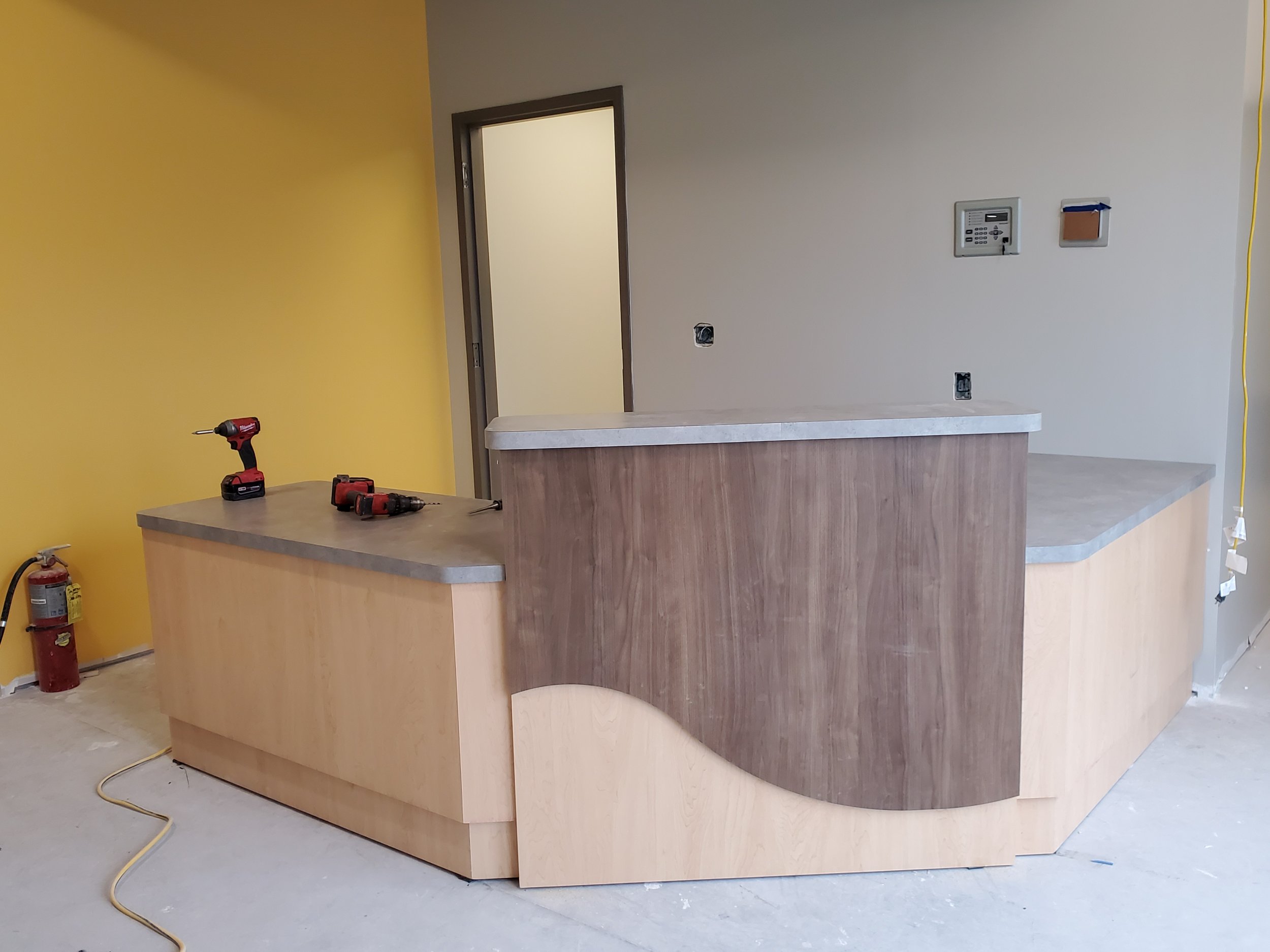 A partially assembled curved wooden reception desk with construction tools on top inside a building under renovation, with yellow painted walls and exposed electrical wires.