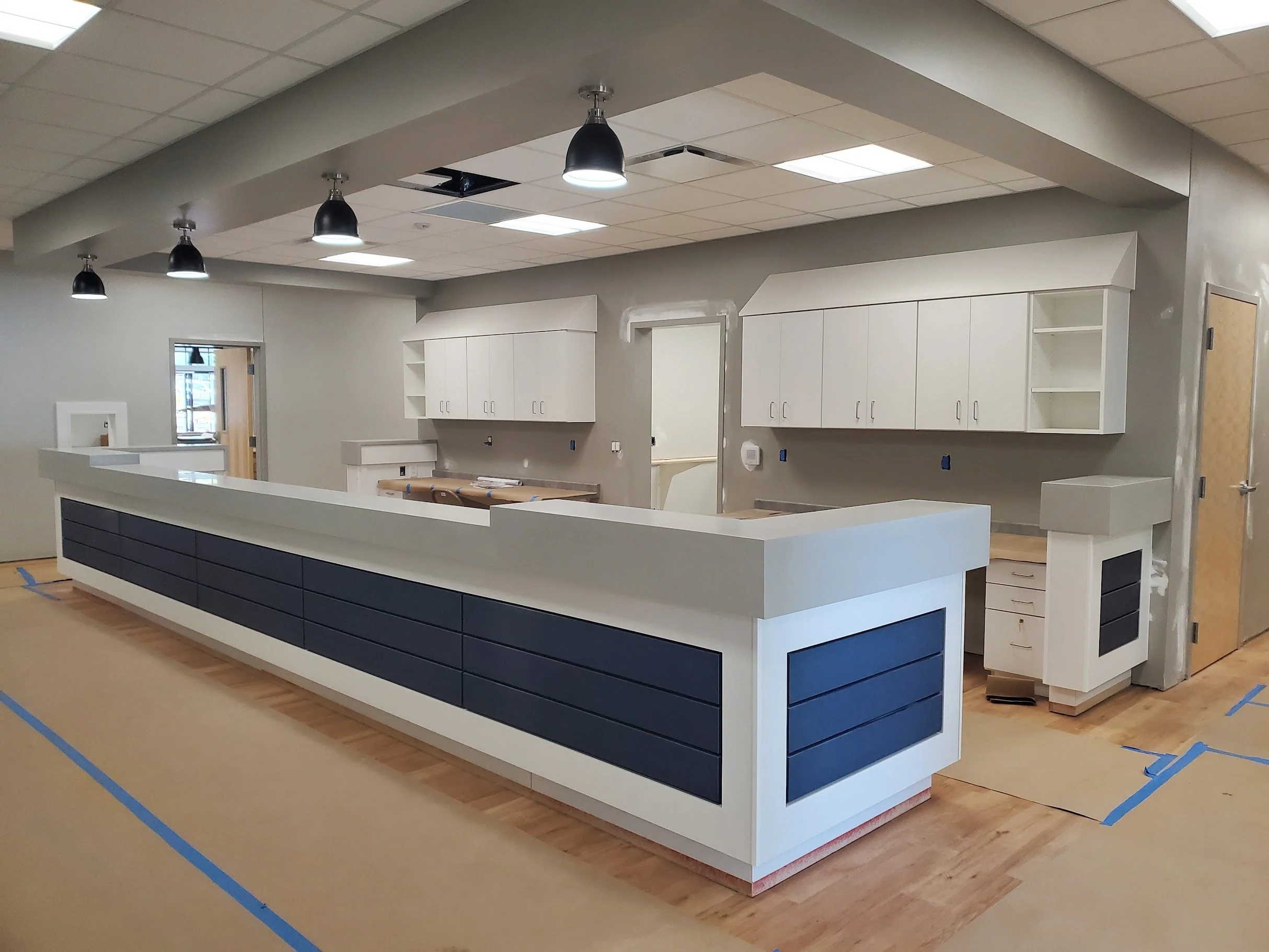 An interior construction site with a large kitchen island, white cabinets, and a suspended ceiling with lighting fixtures. The floors are covered with a protective material and construction markings.