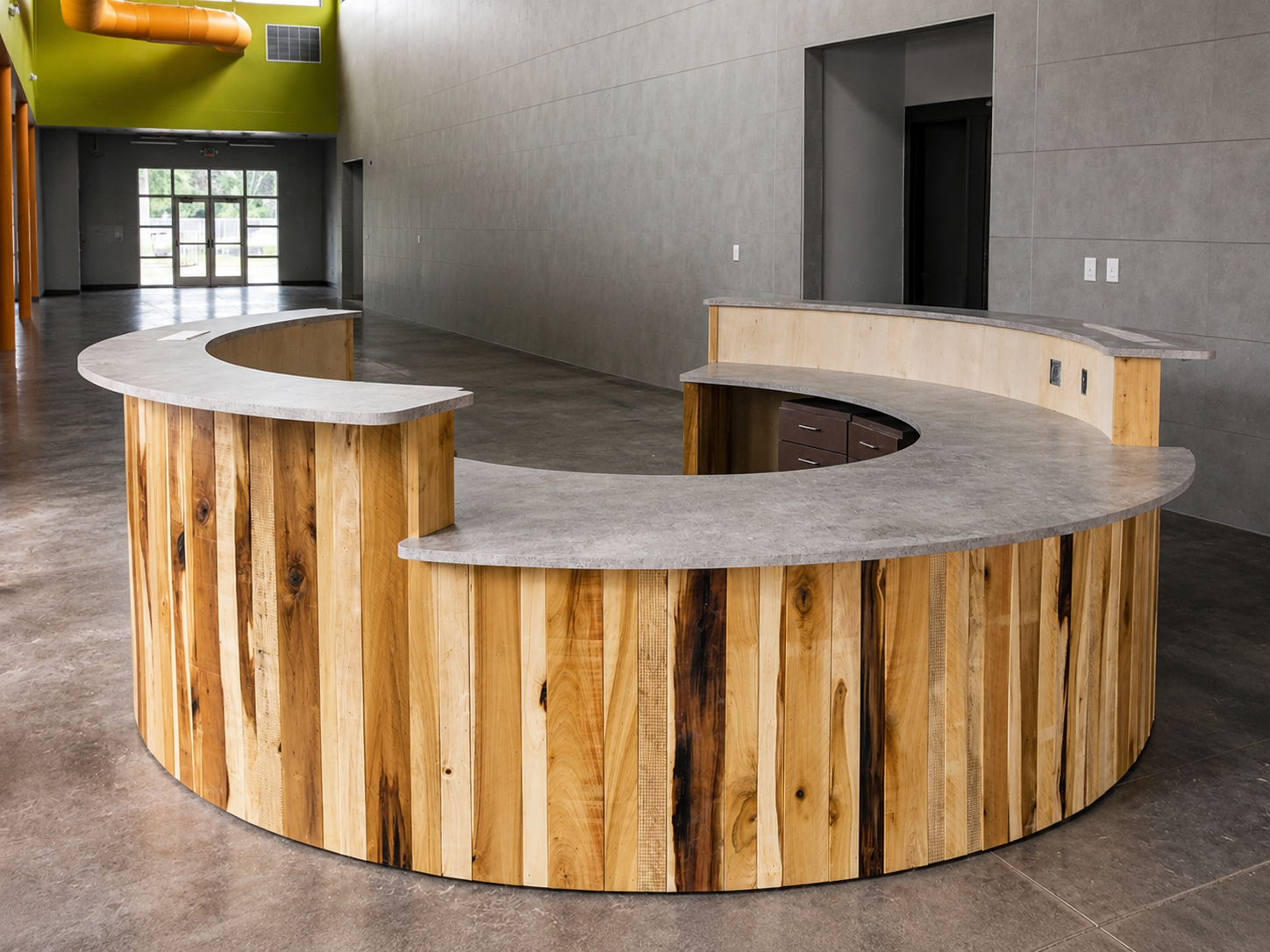 Empty modern reception desk made of wood and concrete in a spacious room with gray walls and large glass doors in the background.