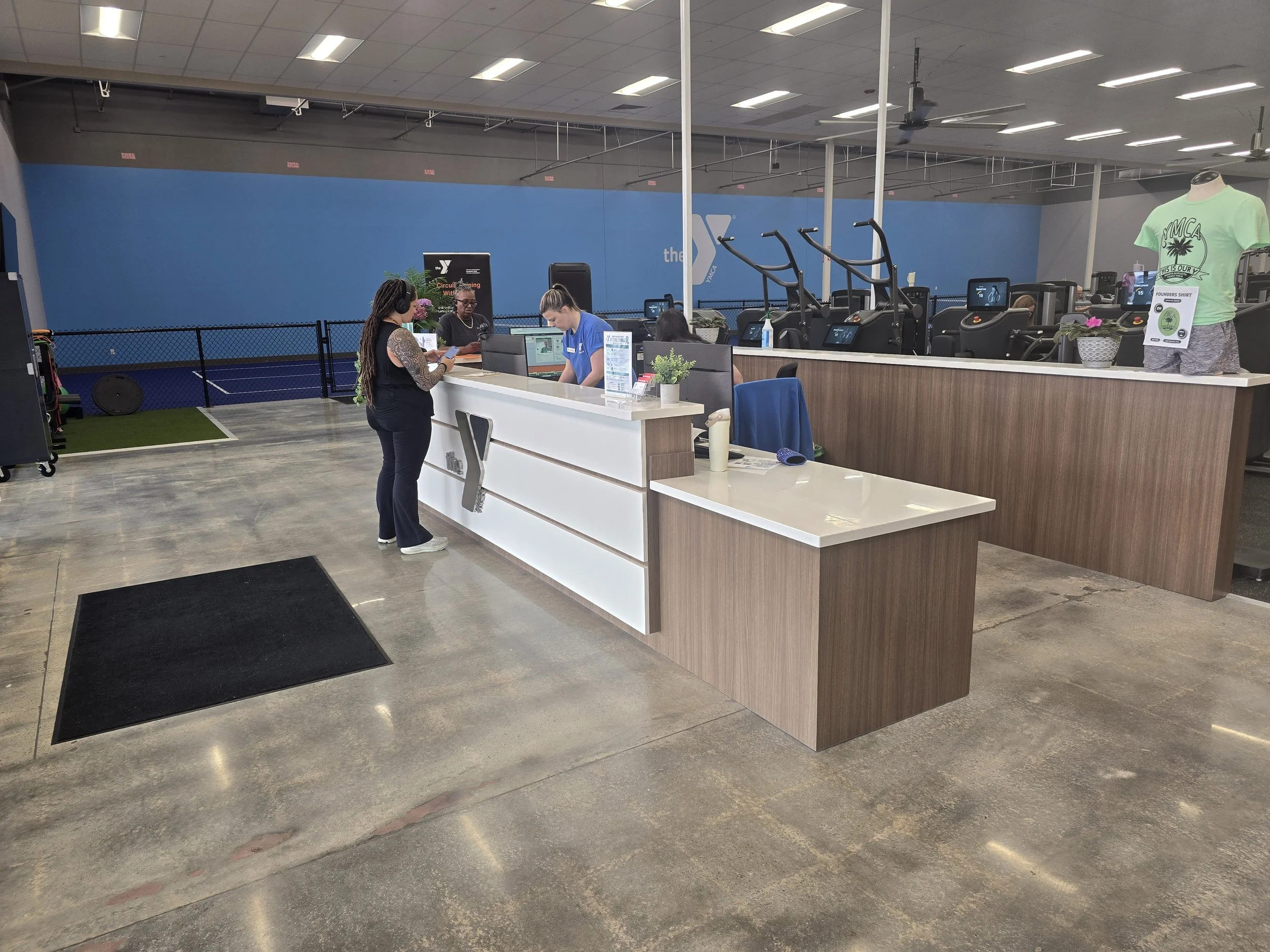 Indoor gym reception area with a white counter, two staff members, and a woman with headphones. Fitness equipment and a mannequin with a YMCA t-shirt are visible in the background.