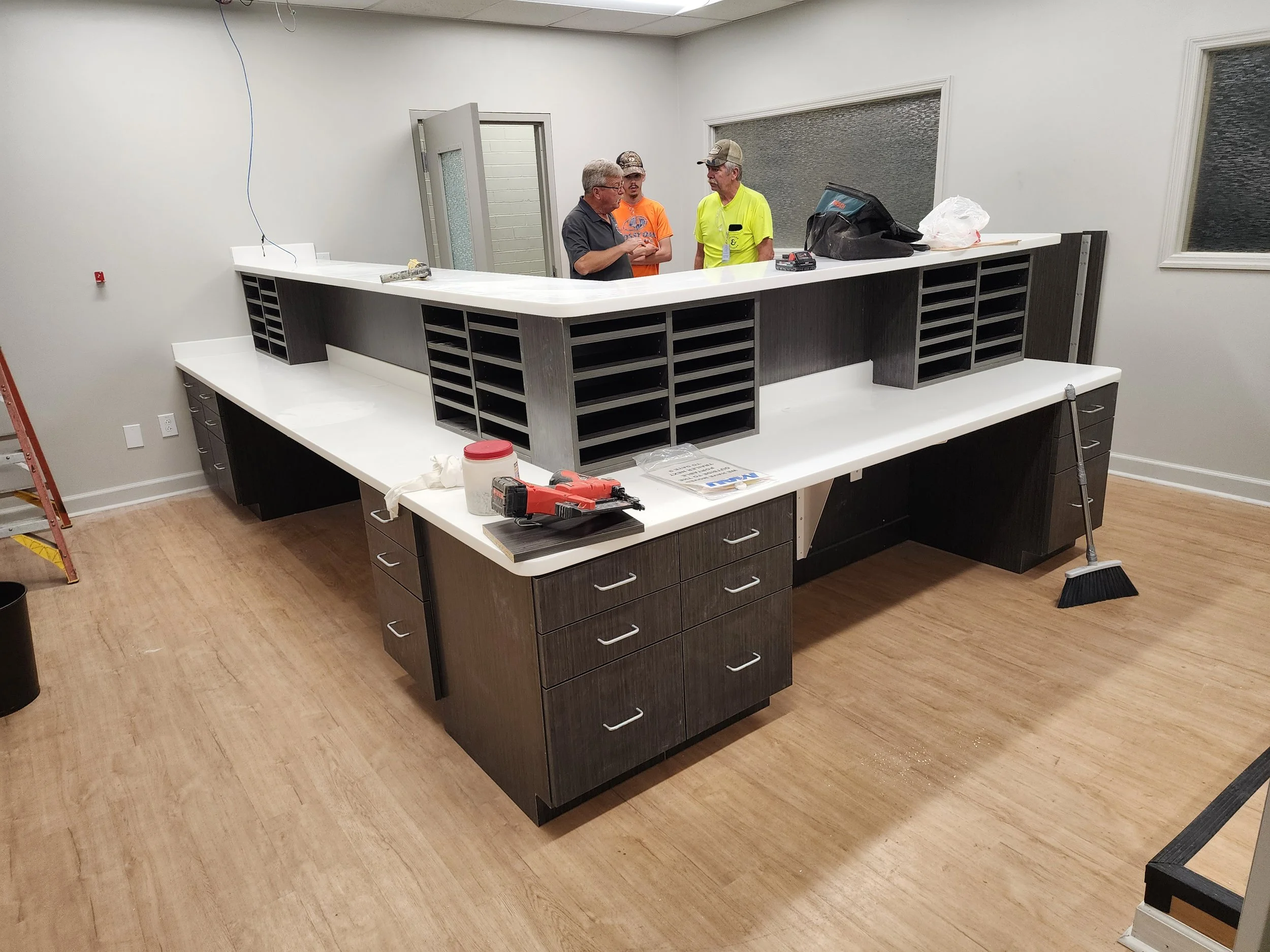 Three men are engaged in a conversation behind a large L-shaped white kitchen island with black cabinetry. One man wears glasses and a dark shirt, the second man is in a bright yellow shirt, and the third is in an orange shirt. The room is under cons