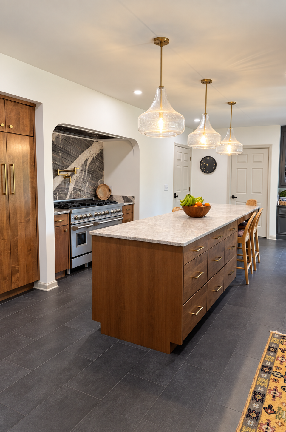 Modern kitchen with a large wooden island, three glass pendant lights, a fruit bowl, and a stainless steel stove with a marble backsplash.