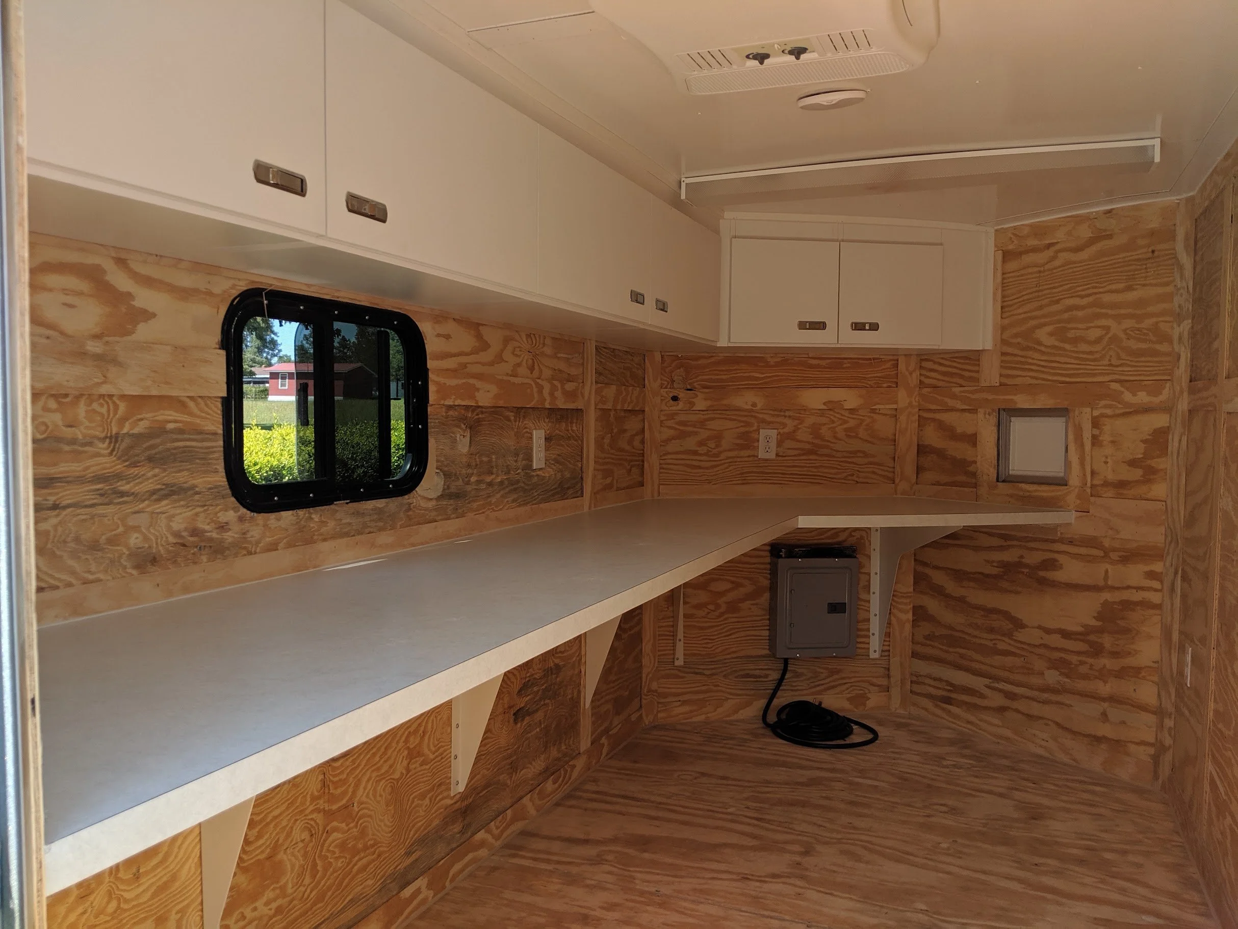Interior of a small wooden space, possibly a trailer or tiny home, with white cabinets overhead, a small window showing an outdoor scene with a red building and greenery, and electrical outlets on the wooden wall.