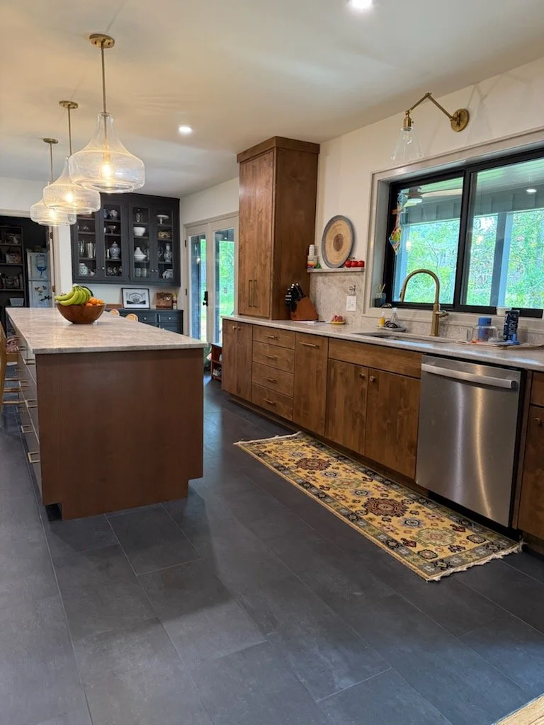 Modern kitchen with wooden cabinets, black glass-front cabinets, granite countertops, stainless steel dishwasher, and a window overlooking greenery. There are hanging pendant and wall-mounted lights, a multicolored area rug, and a kitchen island with