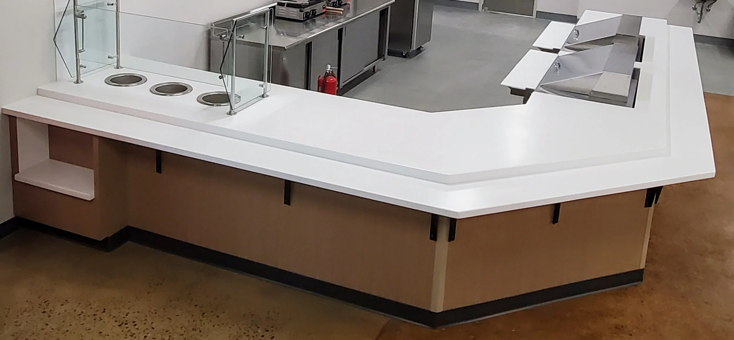 Empty cafeteria or food service counter with a white surface, glass sneeze guard, three circular trays or dishes, and a stainless steel prep area in the background. Fire extinguisher visible behind the counter.