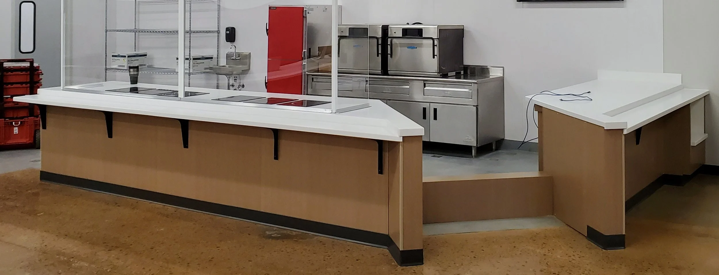 Empty cafeteria serving counter with a plastic shield, stainless steel appliances, and a small prepared food area in the background.