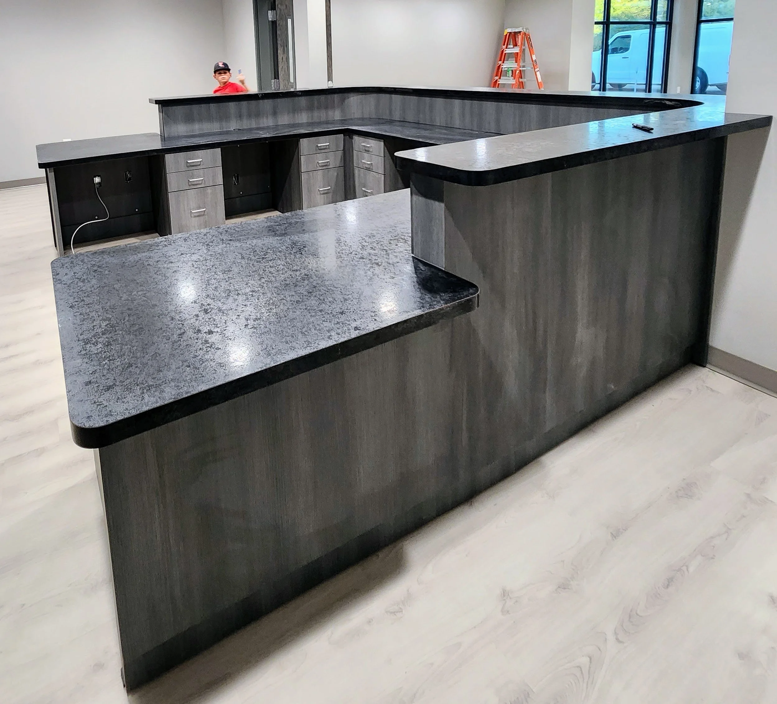 A kitchen island with a black countertop and gray wooden cabinets, with a worker in the background and an orange ladder near a large window.