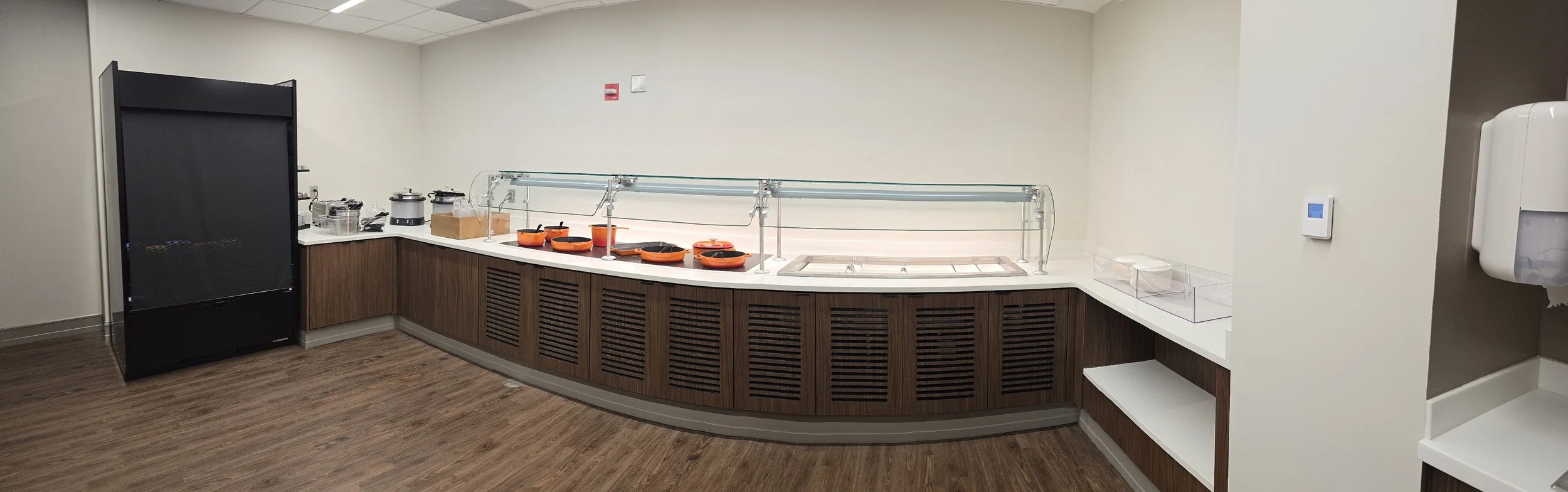 A cafeteria food service area with a black refrigerator on the left, a white counter with various kitchen appliances, and orange bowls with food on a heated serving station. On the right, there is a wall-mounted thermostat and a wall-mounted paper to