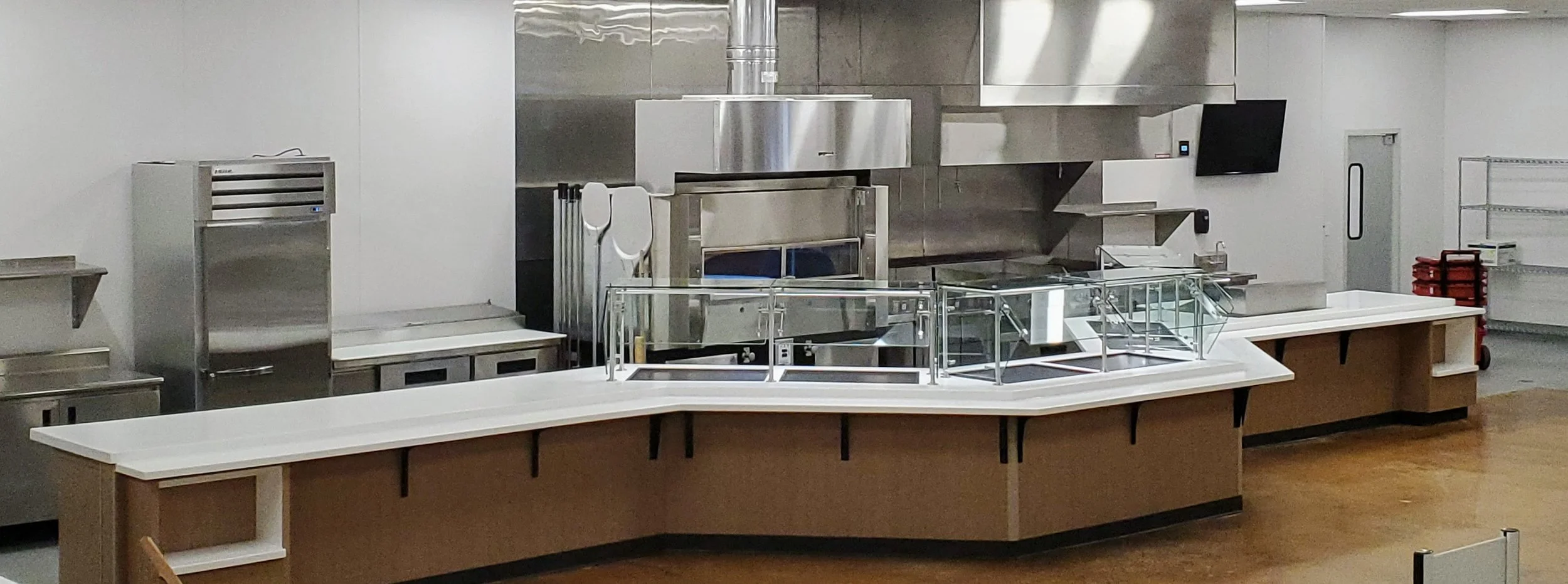 Empty cafeteria serving line with stainless steel appliances, glass display case, and white countertops.