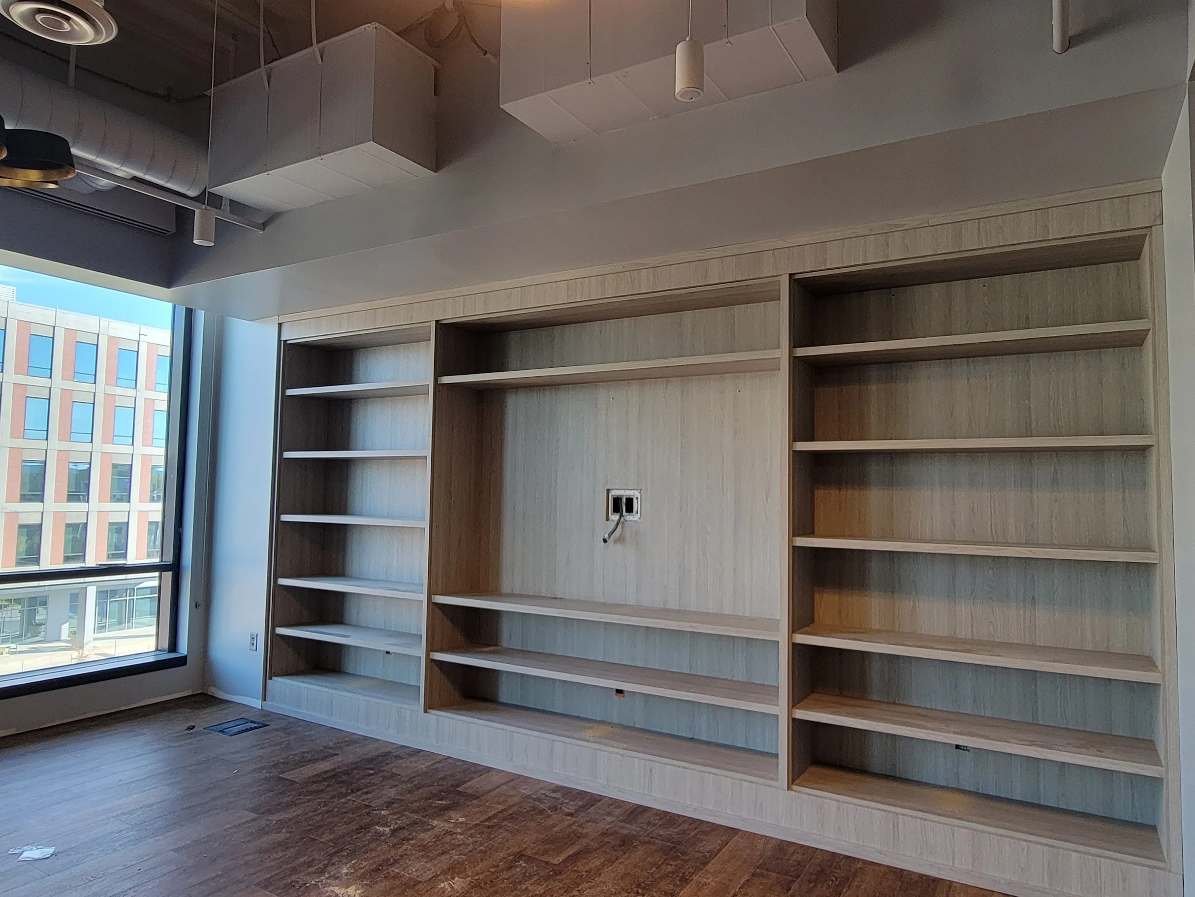 Empty wooden bookshelves in a room with wood flooring and large windows showing a building outside.