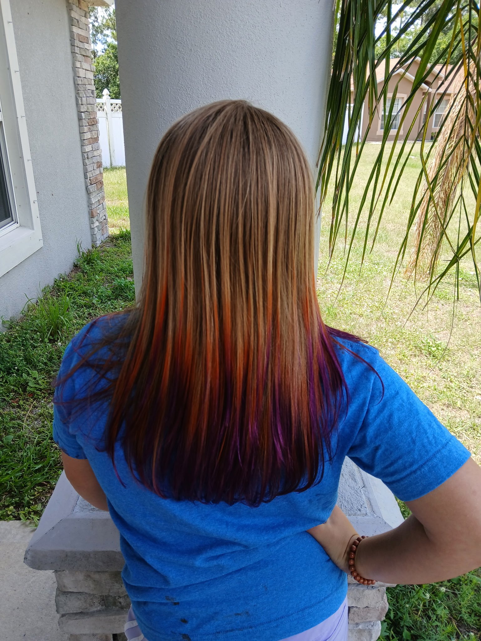 A woman with long, straight, blonde hair streaked with purple, orange, and brown, sitting outdoors on a concrete bench near a white wall with a window, surrounded by green grass, plants, and trees, with her arm resting on a white surface and wearing 