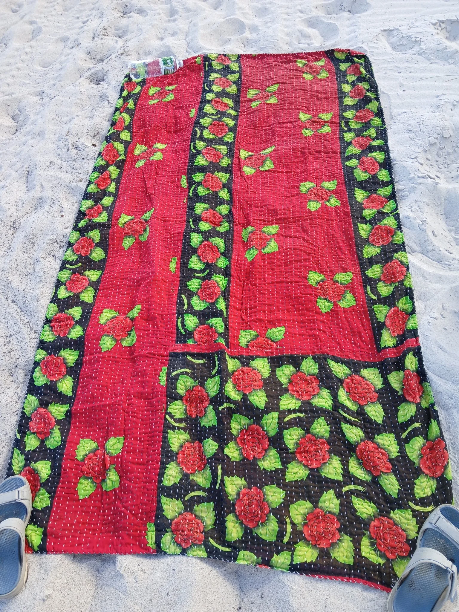A colorful beach towel with red roses and green leaves on a black background, spread out on white sand, with a plastic water bottle at the top and two pairs of shoes at the bottom edges.
