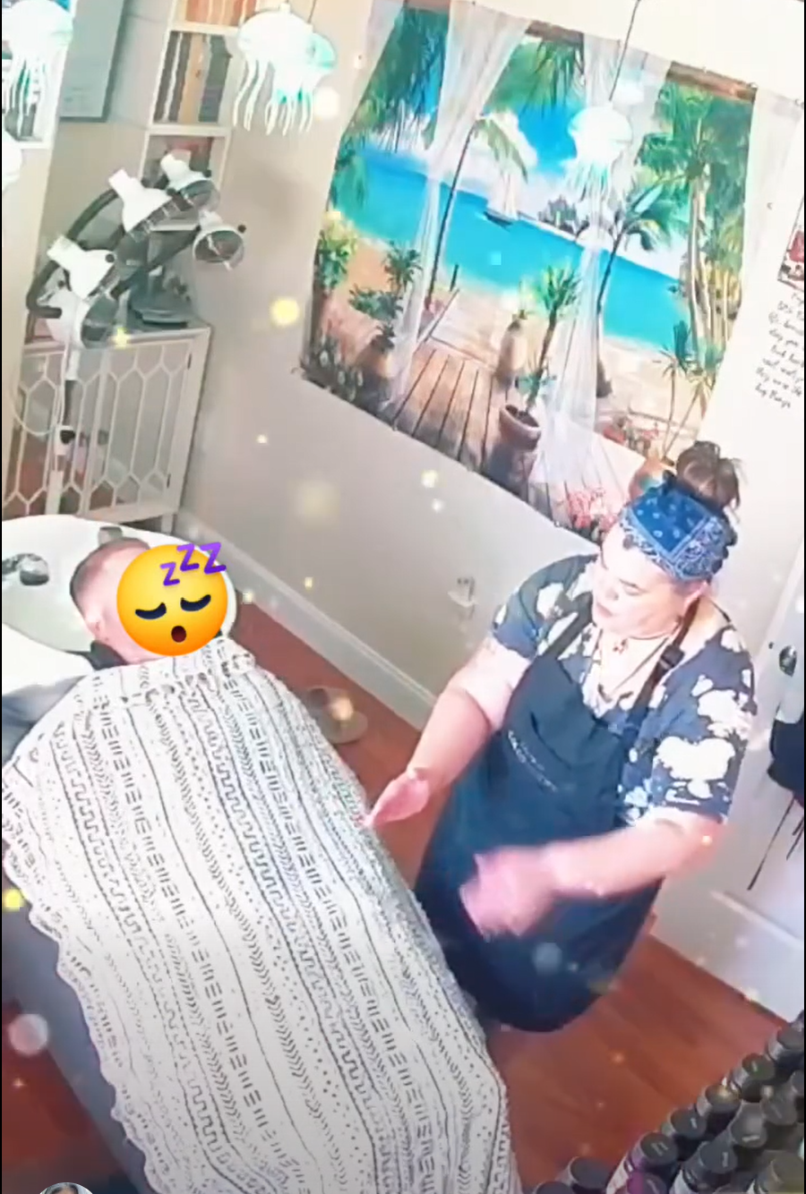 A woman with a blue bandana and floral shirt is offering reiki and energy clearing to client during a hair salon shampoo. 