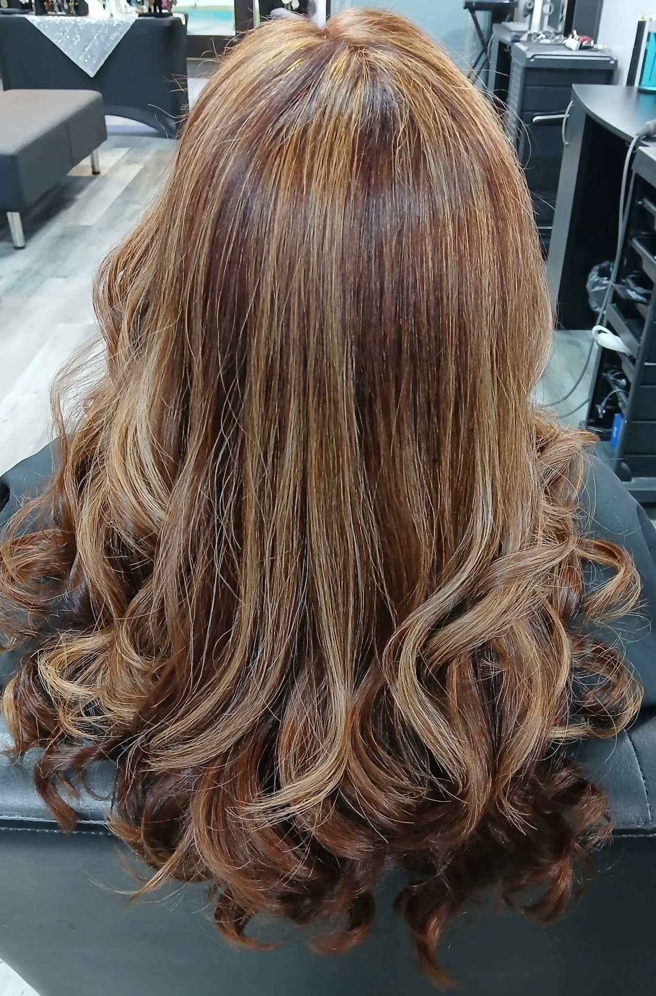 Back view of a woman with long, wavy hair. The hair is dyed in shades of light brown, copper, and auburn with highlights, styled in loose curls. The setting appears to be a salon with chairs and equipment visible in the background.