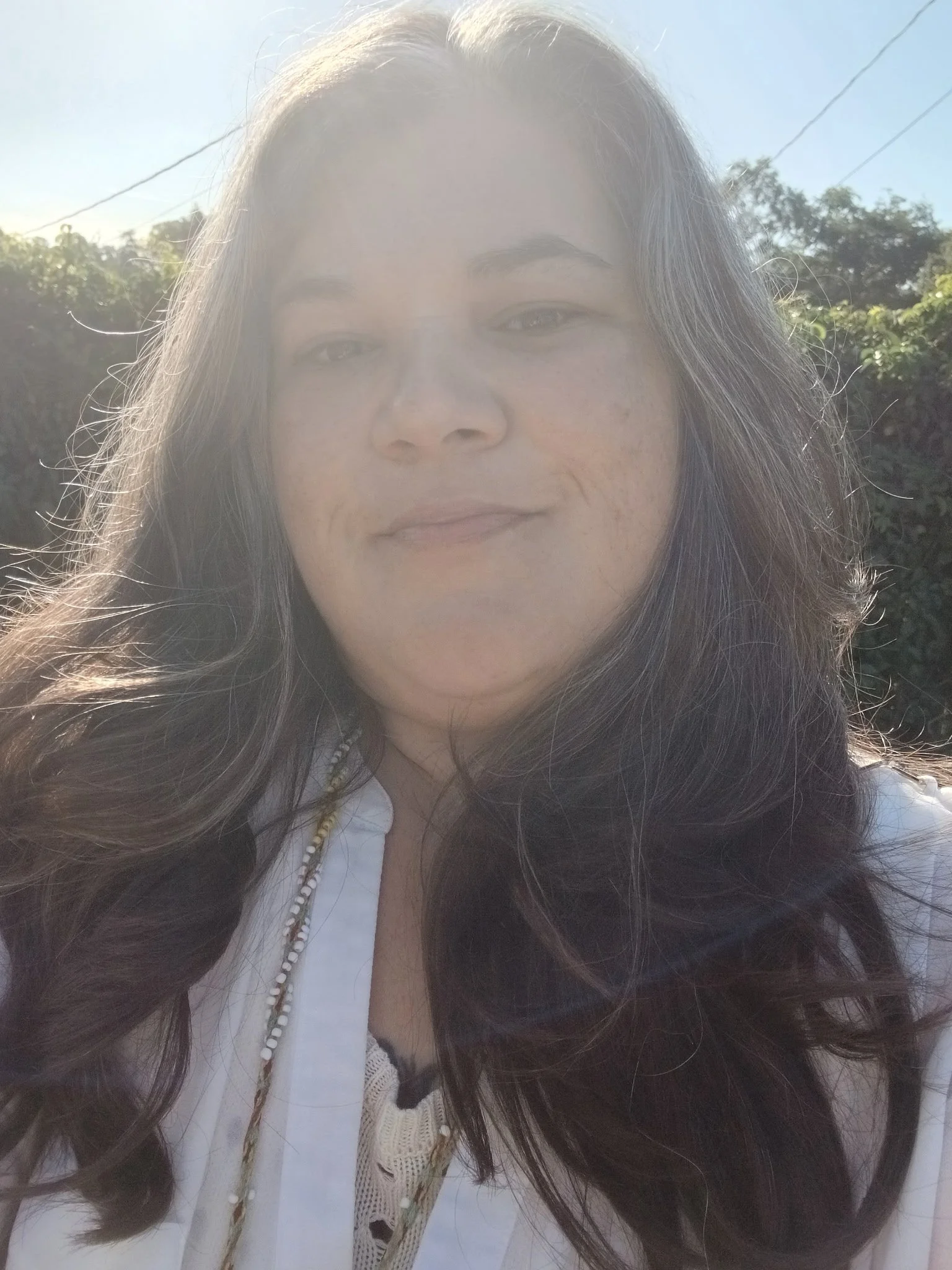 Close-up of a woman with long dark hair, outdoors with sunlight, trees, and clear sky in the background.