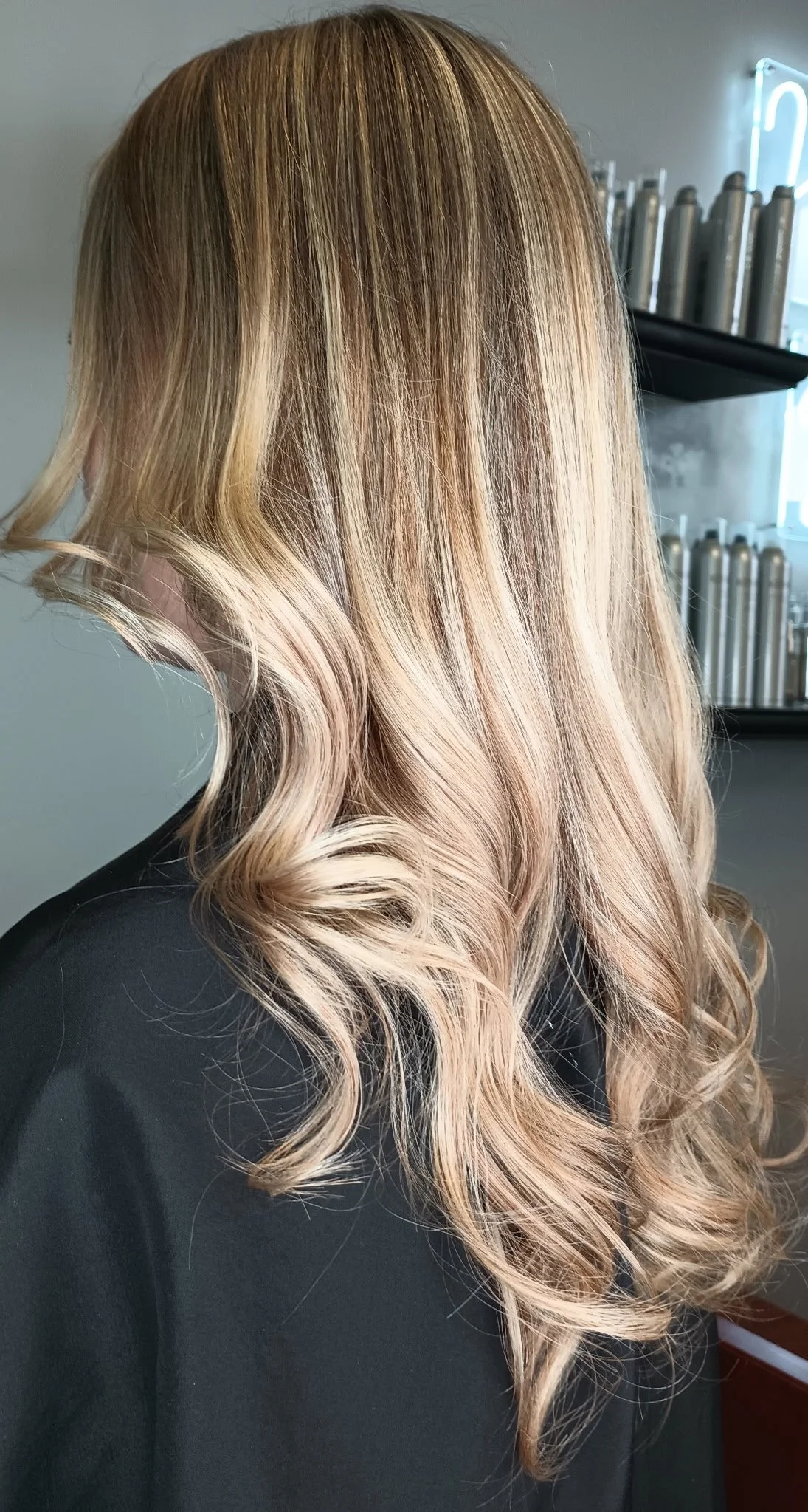 A woman with long, wavy, blonde hair wearing a black top, sitting in a salon with hair products on shelves in the background.