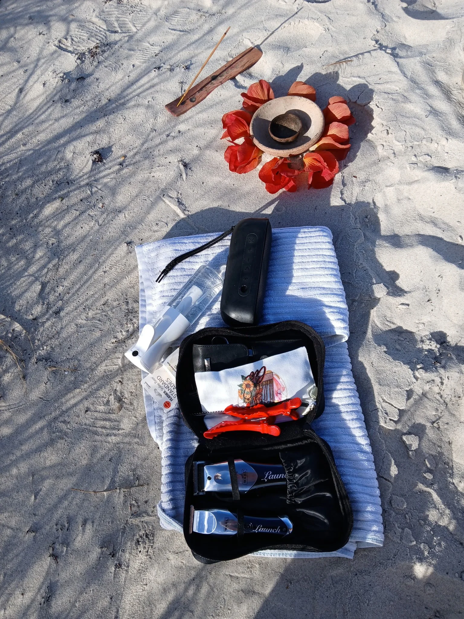 Beach scene with a white towel on the sand, containing a black toiletry bag with bottles, a hairbrush, and orange hair clips. In the background, there is a small wooden object, a stone with a small black bowl, seaweed on the sandy ground. ceremony