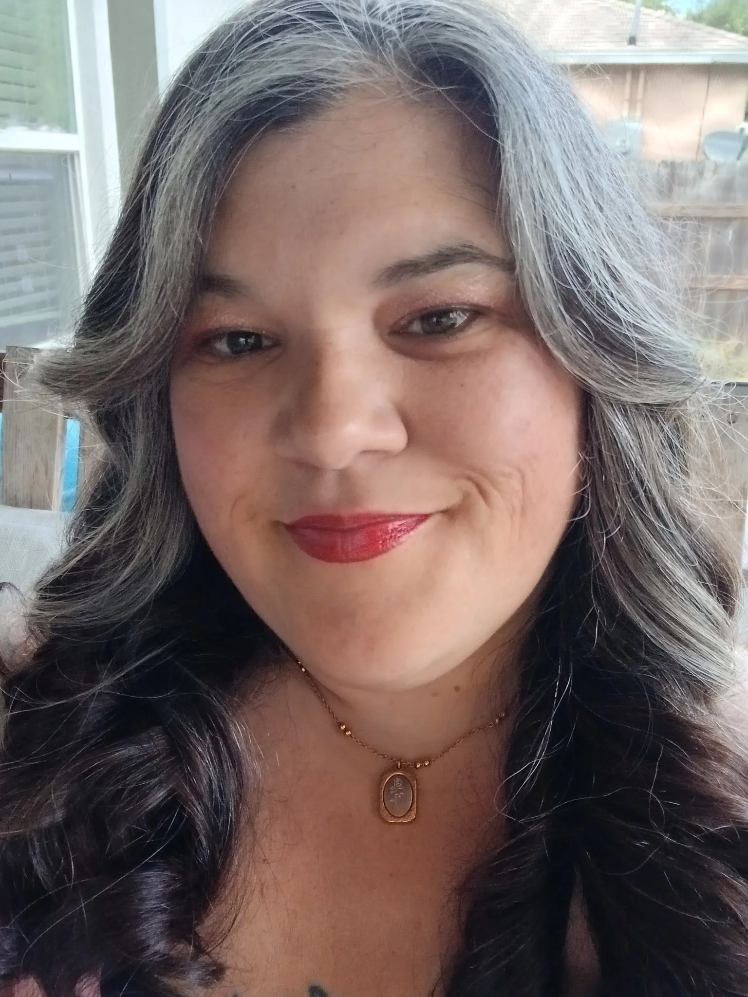 Close-up of a smiling woman with long, wavy gray hair, wearing a gold necklace with a pendant, indoors with a window and a wooden fence in the background.