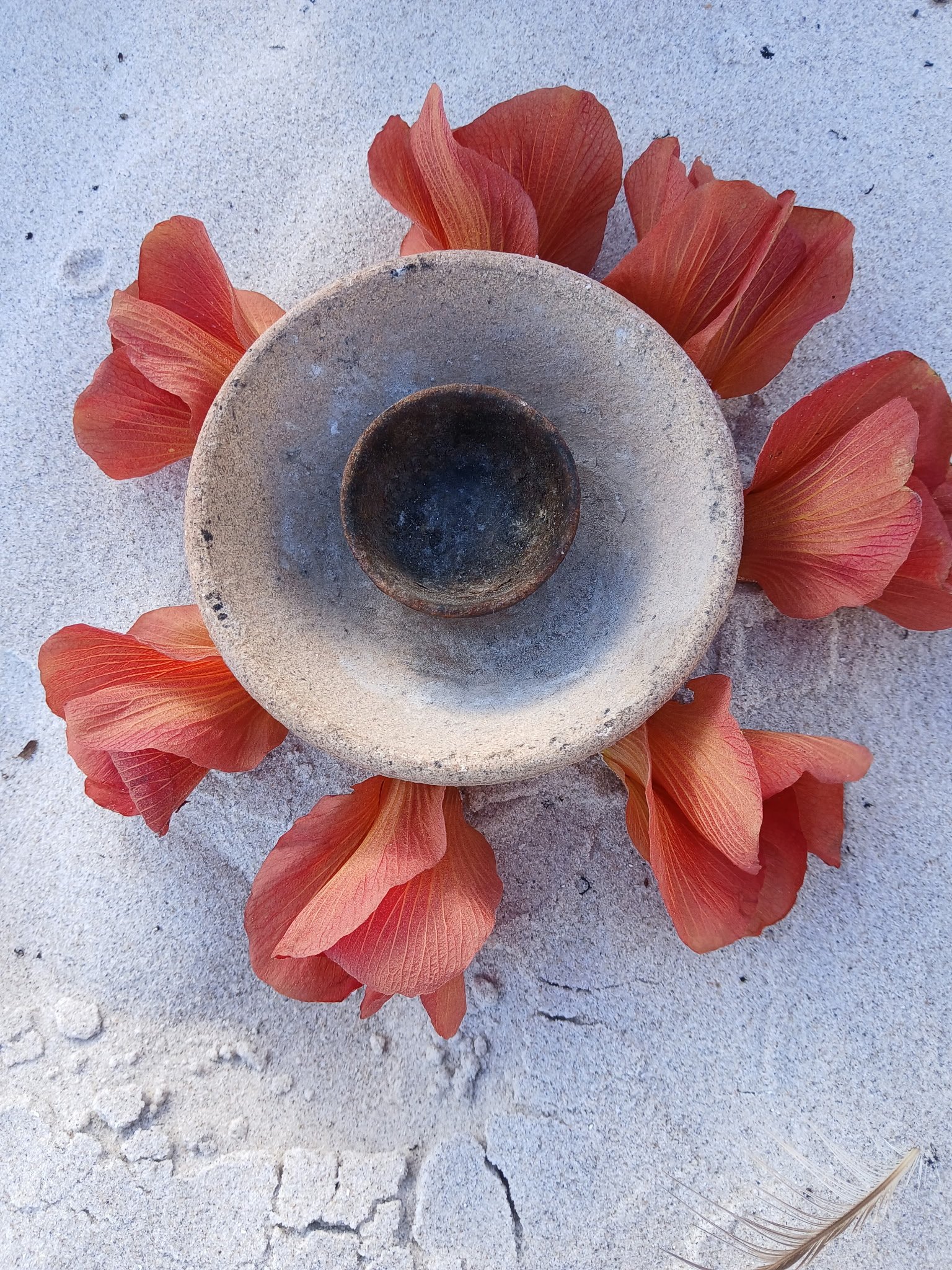 A concrete candle holder surrounded by orange and red flower petals on sandy ground with a feather in the lower right corner.