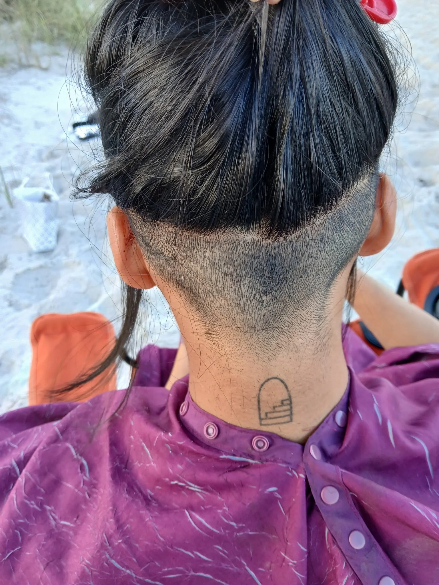 Close-up of a person's head showing a unique haircut with a shaved sides and long black hair on top, wearing a purple tie-dye shirt, outside on a sandy area.