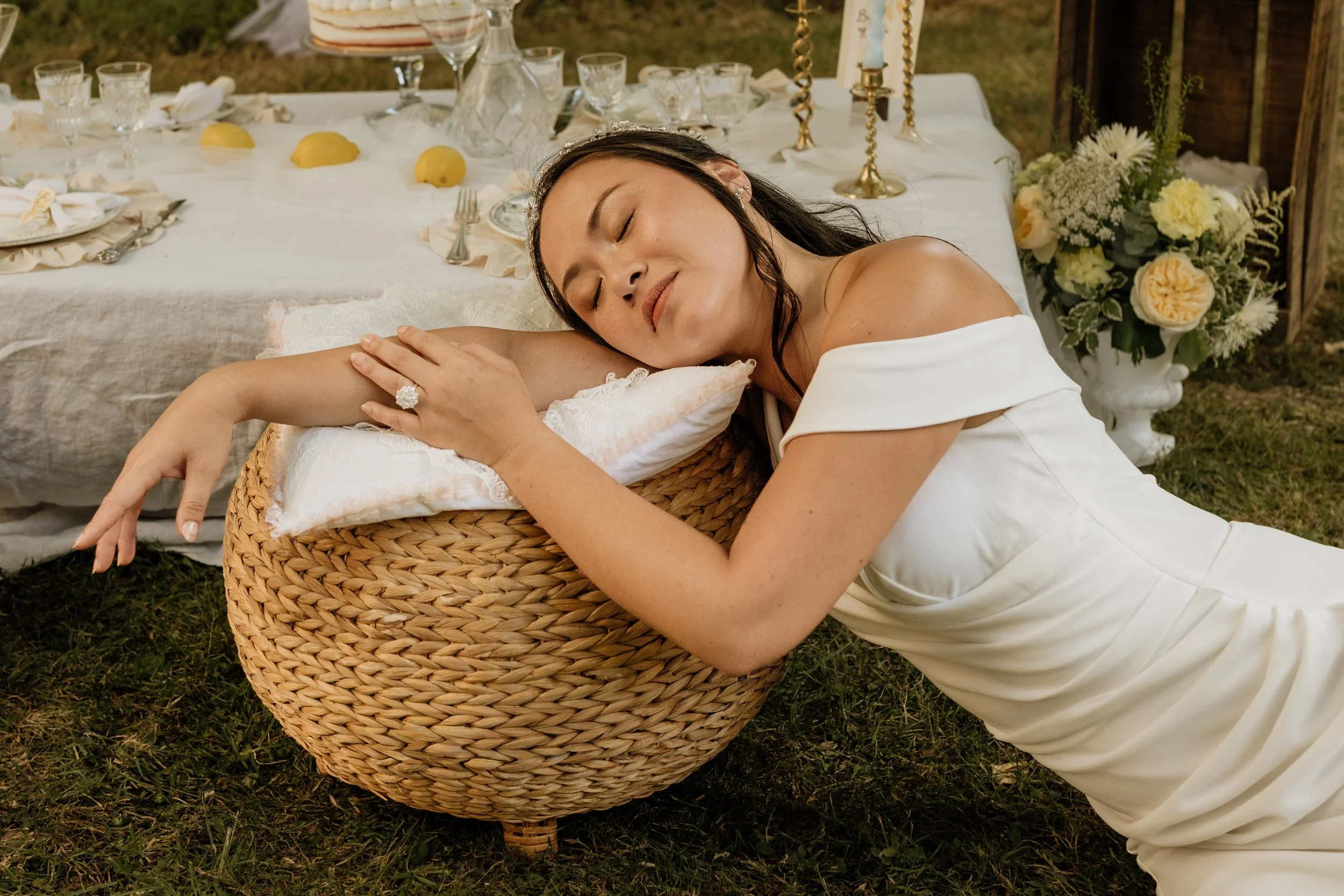 Une femme en robe blanche repose avec les yeux fermés dans un panier en osier, à côté d'une table dressée pour un repas en plein air, avec des fleurs et des décorations à Rennes
