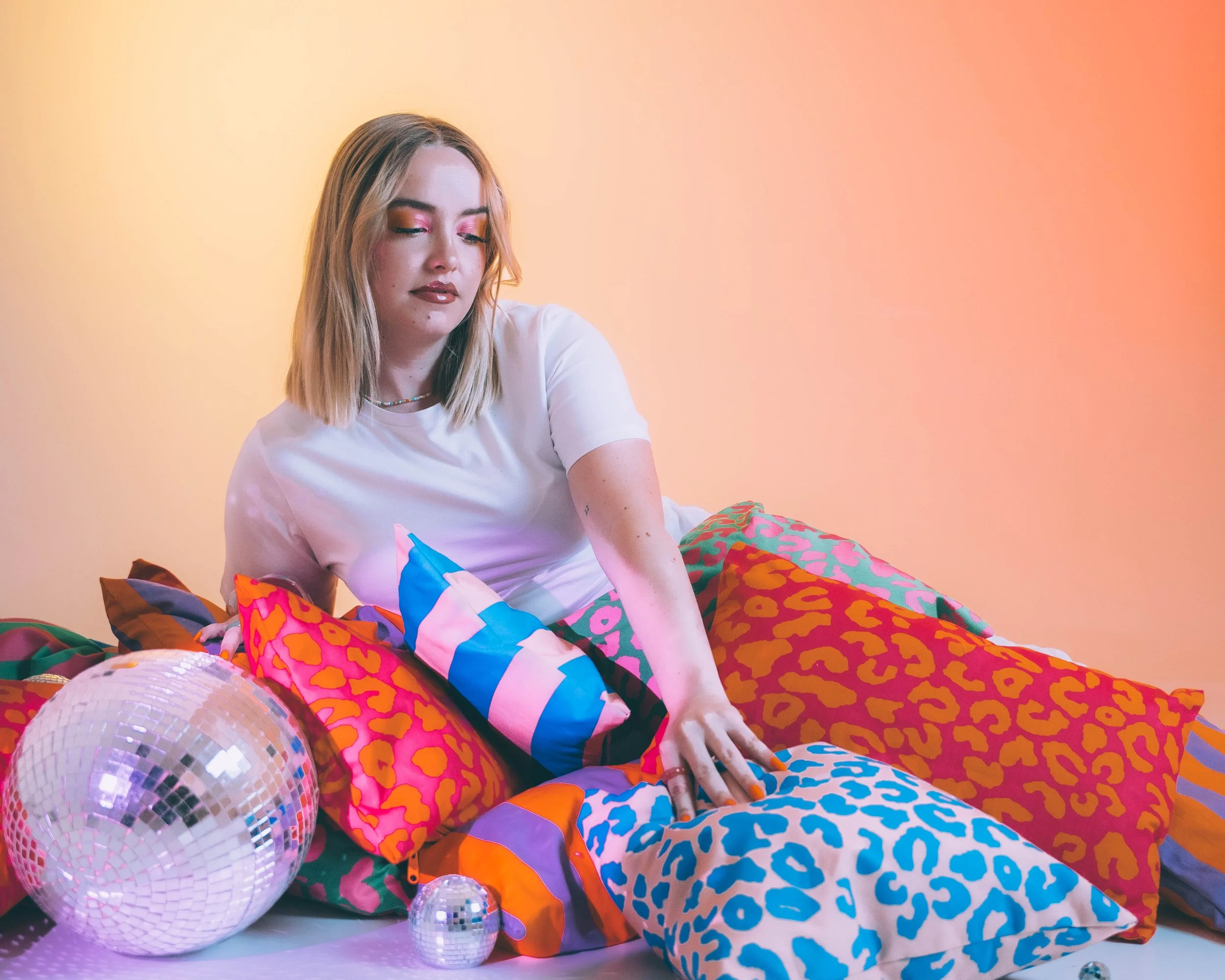 Une jeune femme avec des lèvres foncées et maquillage coloré, assise sur un sol avec des décorations en peluche et ballons, devant un fond dégradé jaune-orange.