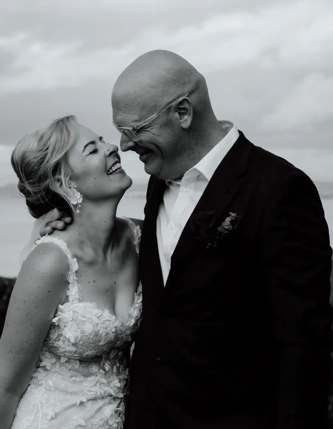 A joyful couple on their wedding day, smiling and looking into each other's eyes, outdoors with a cloudy sky.