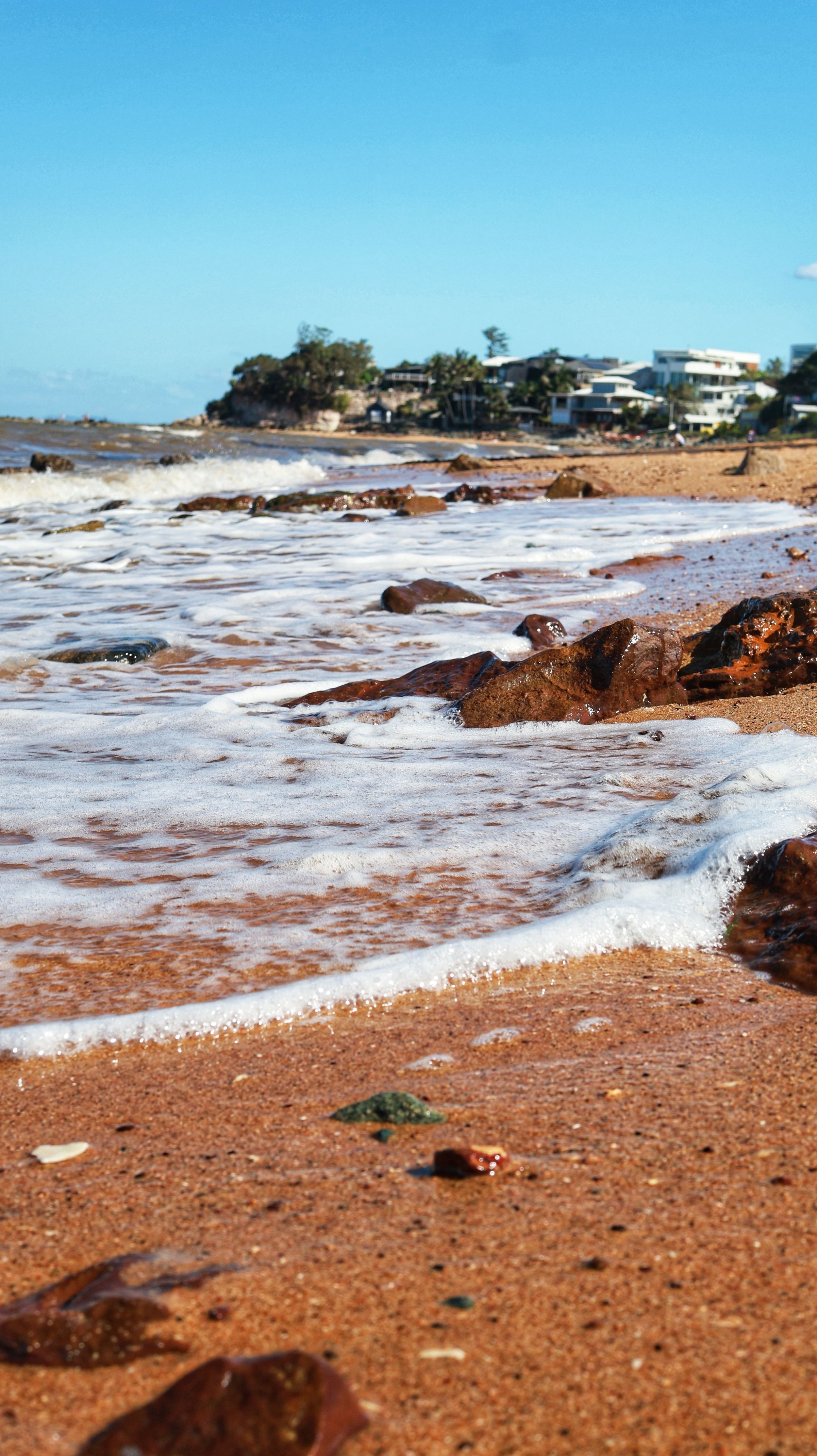View of a sandy beach with rocks in the foreground and a calm ocean with small waves. In the background, there are modern houses on a small hill with trees and a clear blue sky.