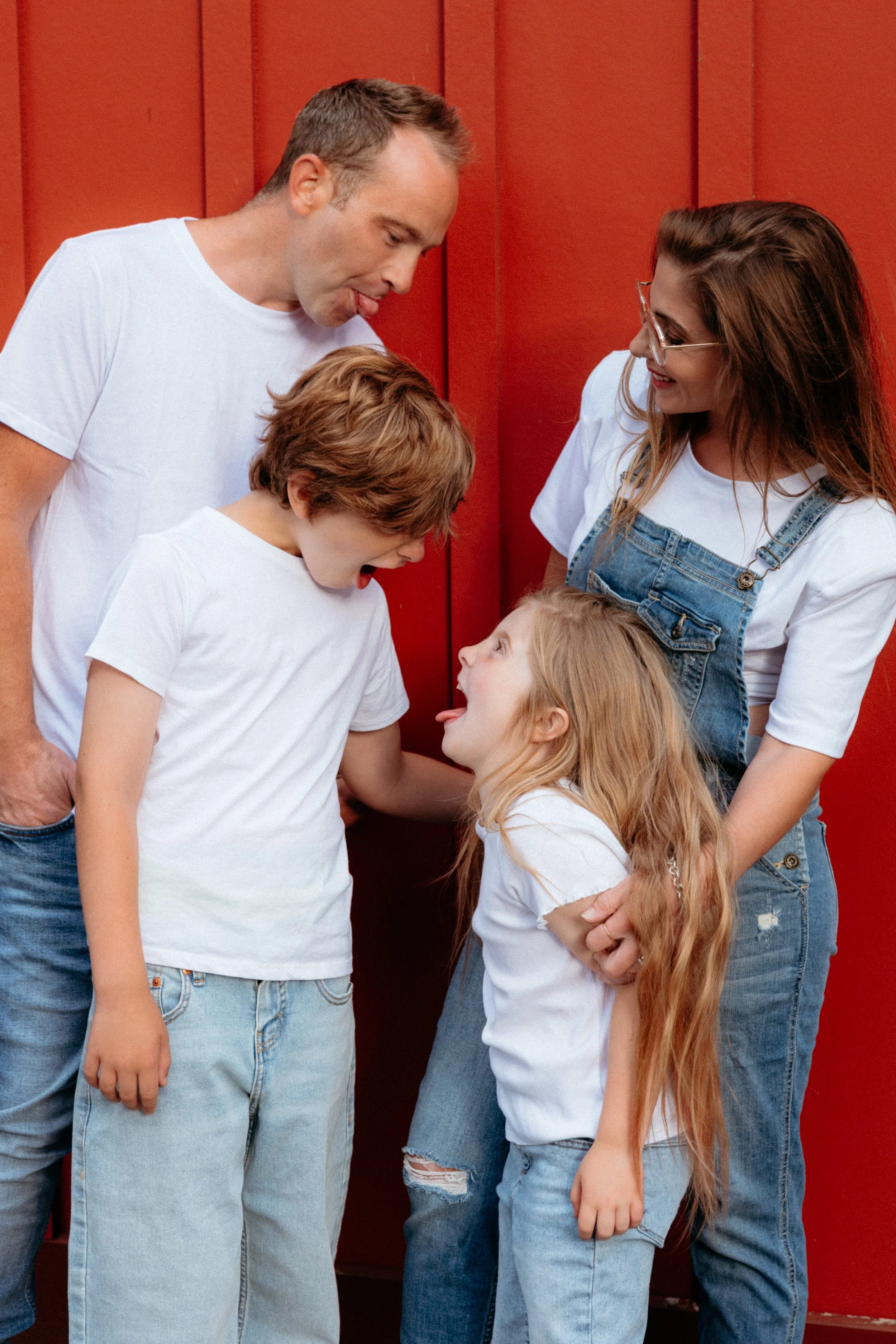 A family of five with two adults and three children interacting playfully against a red background. The man, woman, and children are all smiling and making funny faces.