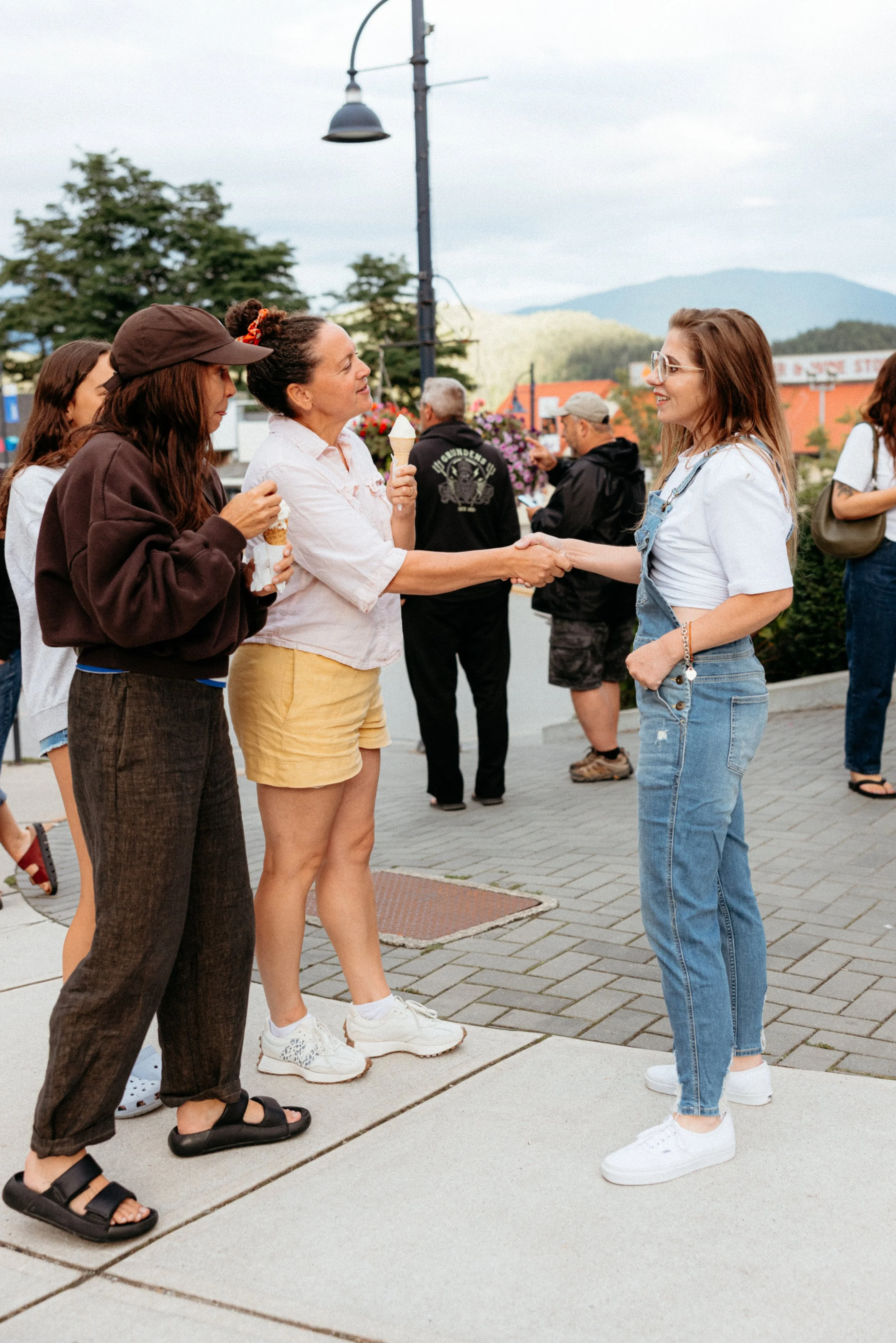 Two women shaking hands on a sidewalk, one holding an ice cream cone, with several people and trees in the background.