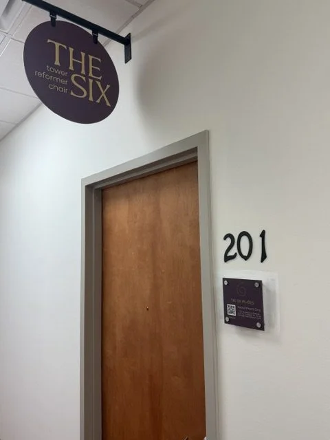A wooden door marked with the number 201, beside a sign with a QR code and text, under a purple sign hanging from the ceiling that reads "THE SIX, tower former chair."