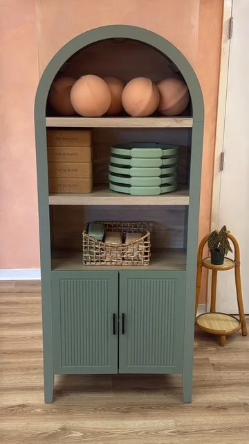 A light green arched cabinet with two doors at the bottom, open middle shelves with stacked pottery, trays, and a wicker basket, set against a peach wall with a small yellow chair and potted plant to the side.
