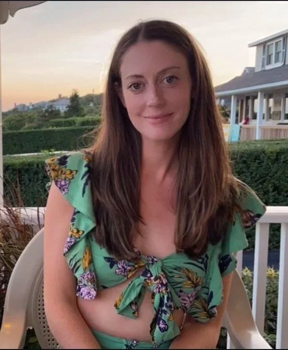 A young woman with long brown hair, wearing a green floral top with ruffled sleeves and a front tie, smiling outdoors on a patio during sunset.