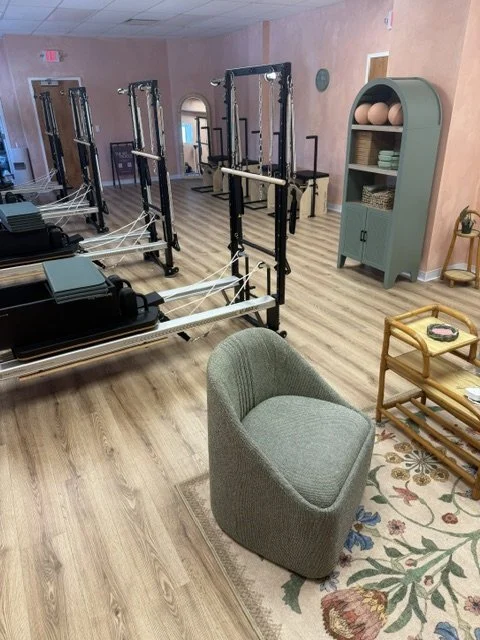 Pilates studio with reformer machines, a green cushioned chair, a teal storage cabinet with gym balls and bowls, and a small wooden table with magazines on a floral rug.