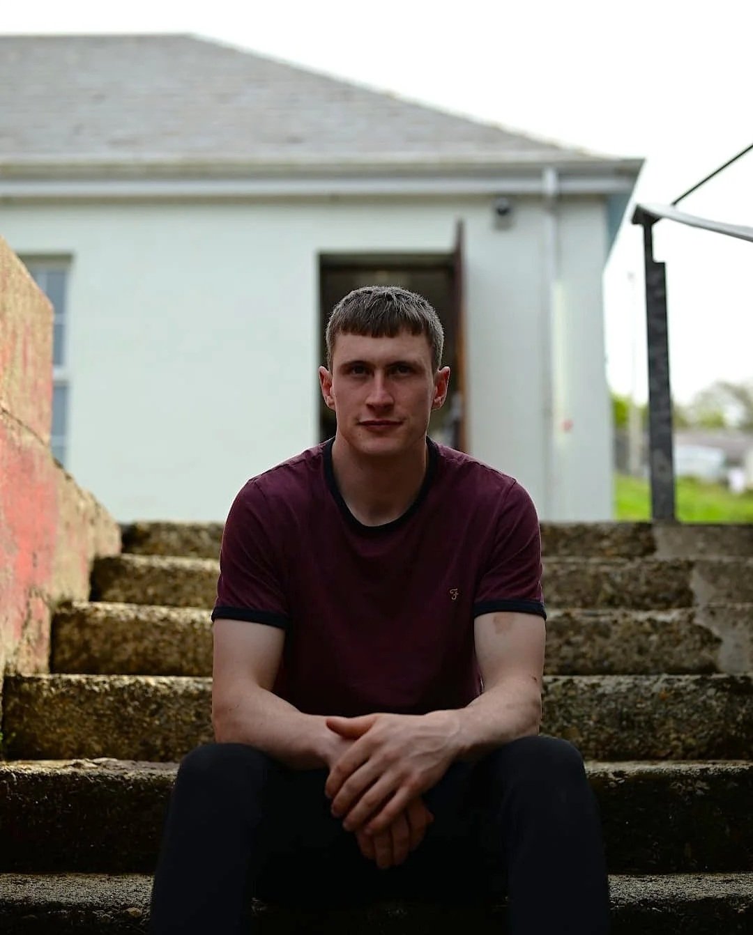 A young man with short brown hair, wearing a maroon t-shirt and black pants, sitting on outdoor stone steps in front of a white building with an open door.