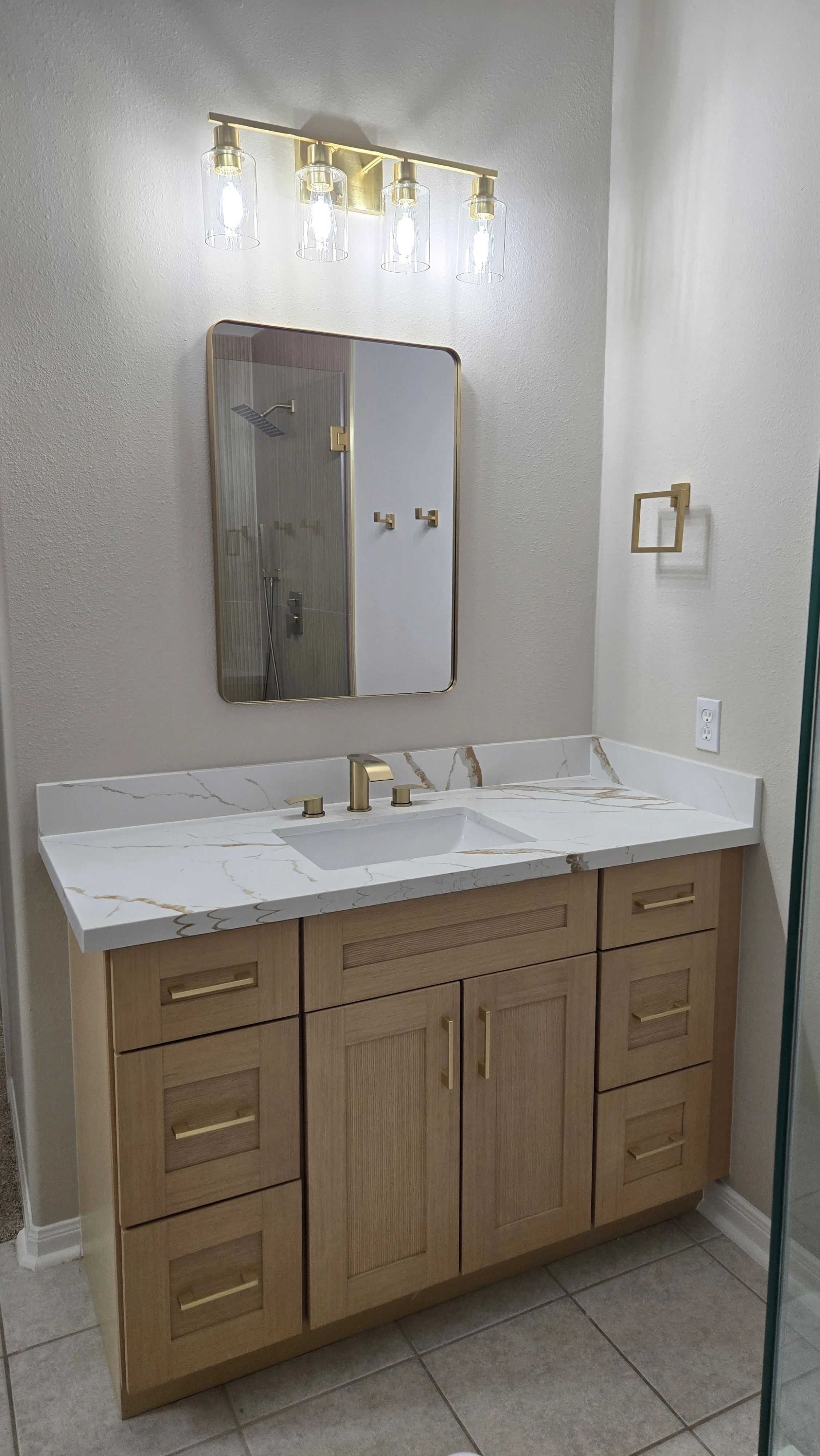 TurnKey Roofing & Restoration — A newly renovated / remodeled bathroom vanity with light wood cabinet, white marble countertop, and gold fixtures, mirror, and towel ring. Houston, Texas