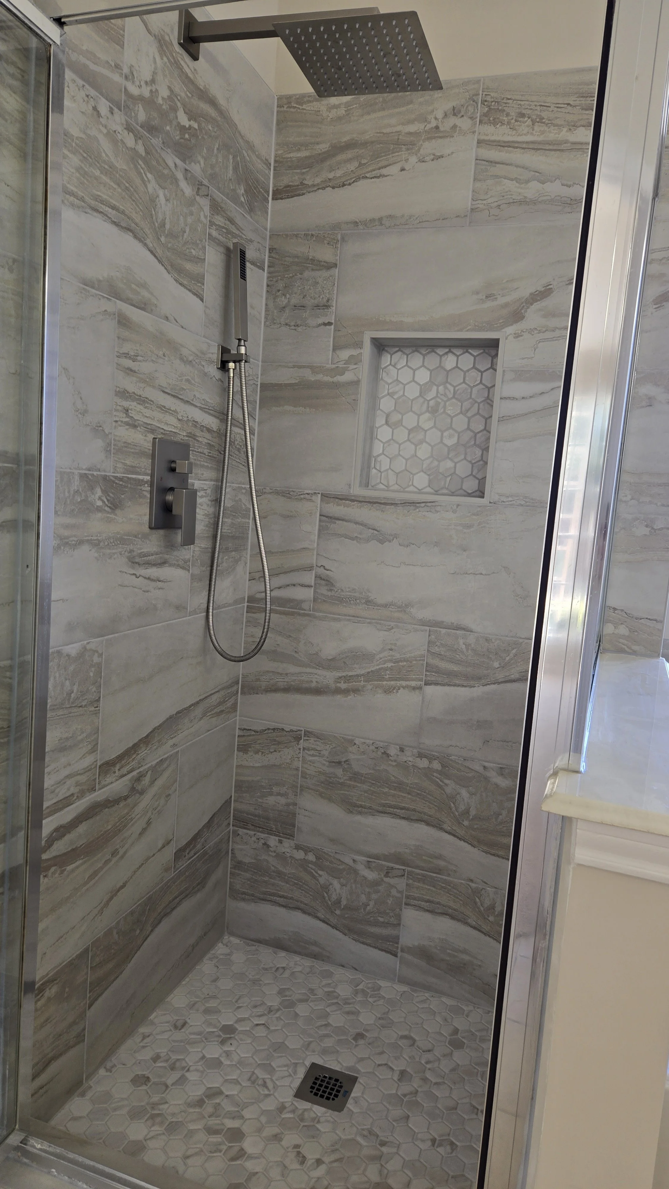 TurnKey Roofing & Restoration — A newly renovated / remodeled modern walk-in shower with large marble-look tiles, a built-in hexagonal tile niche, small hexagonal floor tiles, and a high shower head. Houston, Texas