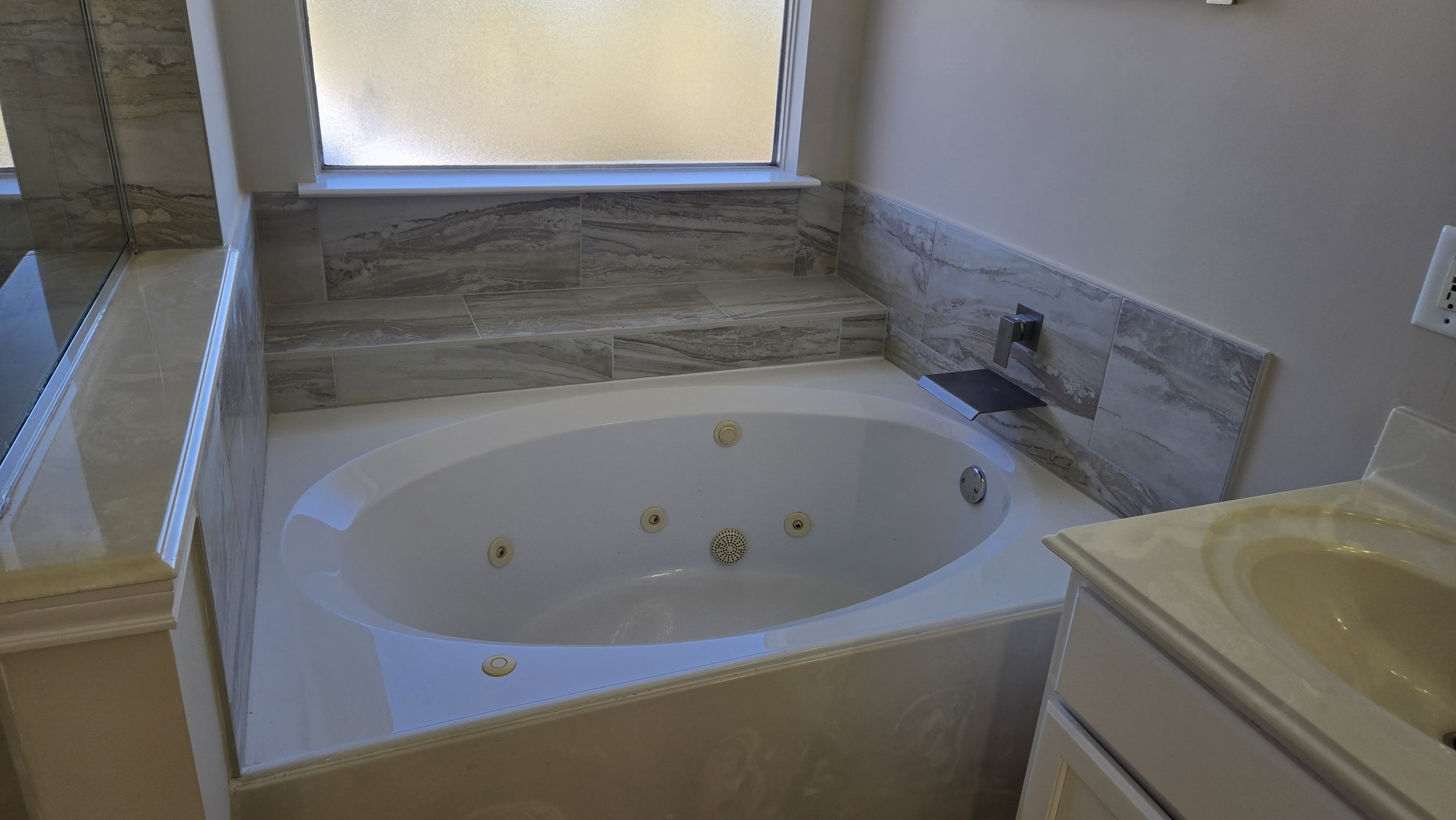 TurnKey Roofing & Restoration — A newly renovated / remodeled bathroom with a white bathtub featuring whirlpool jets, a marble tile surround, a large frosted glass window, a digital control panel on the wall, and a vanity. Houston, Texas