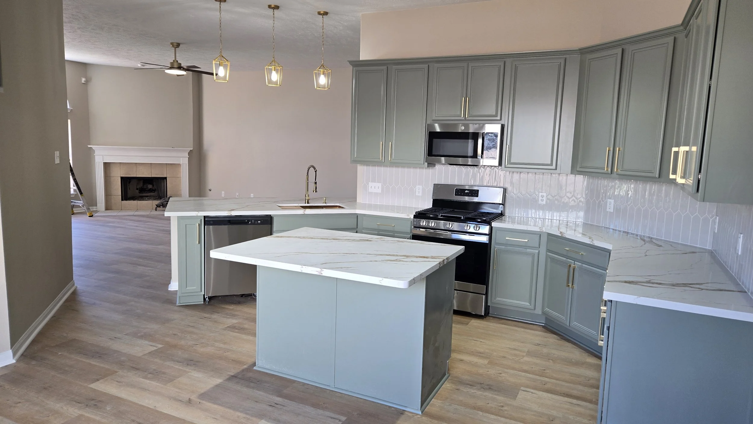 TurnKey Roofing & Restoration — A remodeled modern kitchen with blue green cabinets, white marble countertops, stainless steel appliances, and a kitchen island. In the background, near a fireplace, there are hanging pendant lights and a ceiling fan.