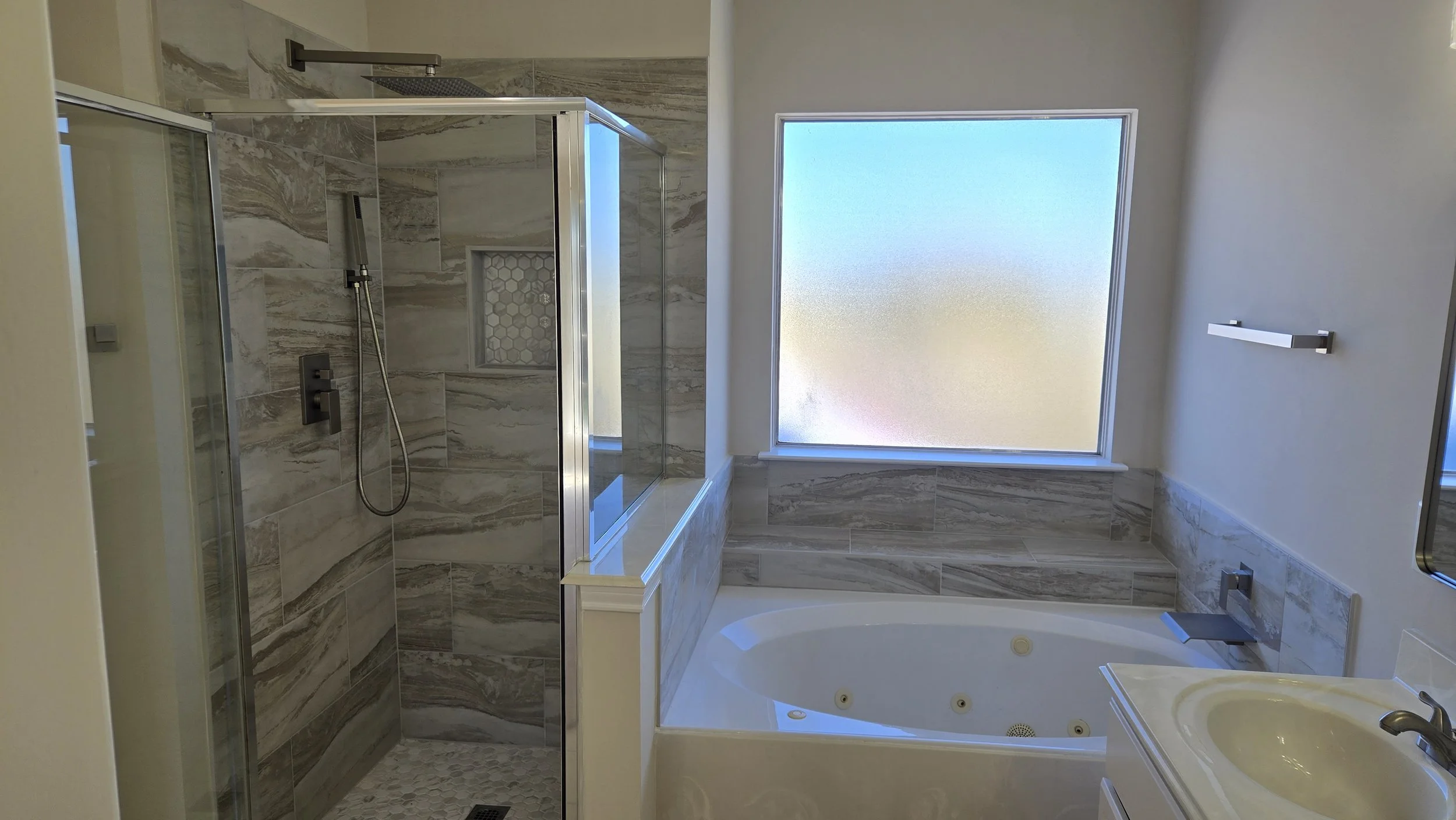 TurnKey Roofing & Restoration — A newly renovated / remodeled modern bathroom featuring a walk-in shower with a glass door, marble tiles, a built-in niche, a frosted window, and a corner bathtub. Houston, Texas