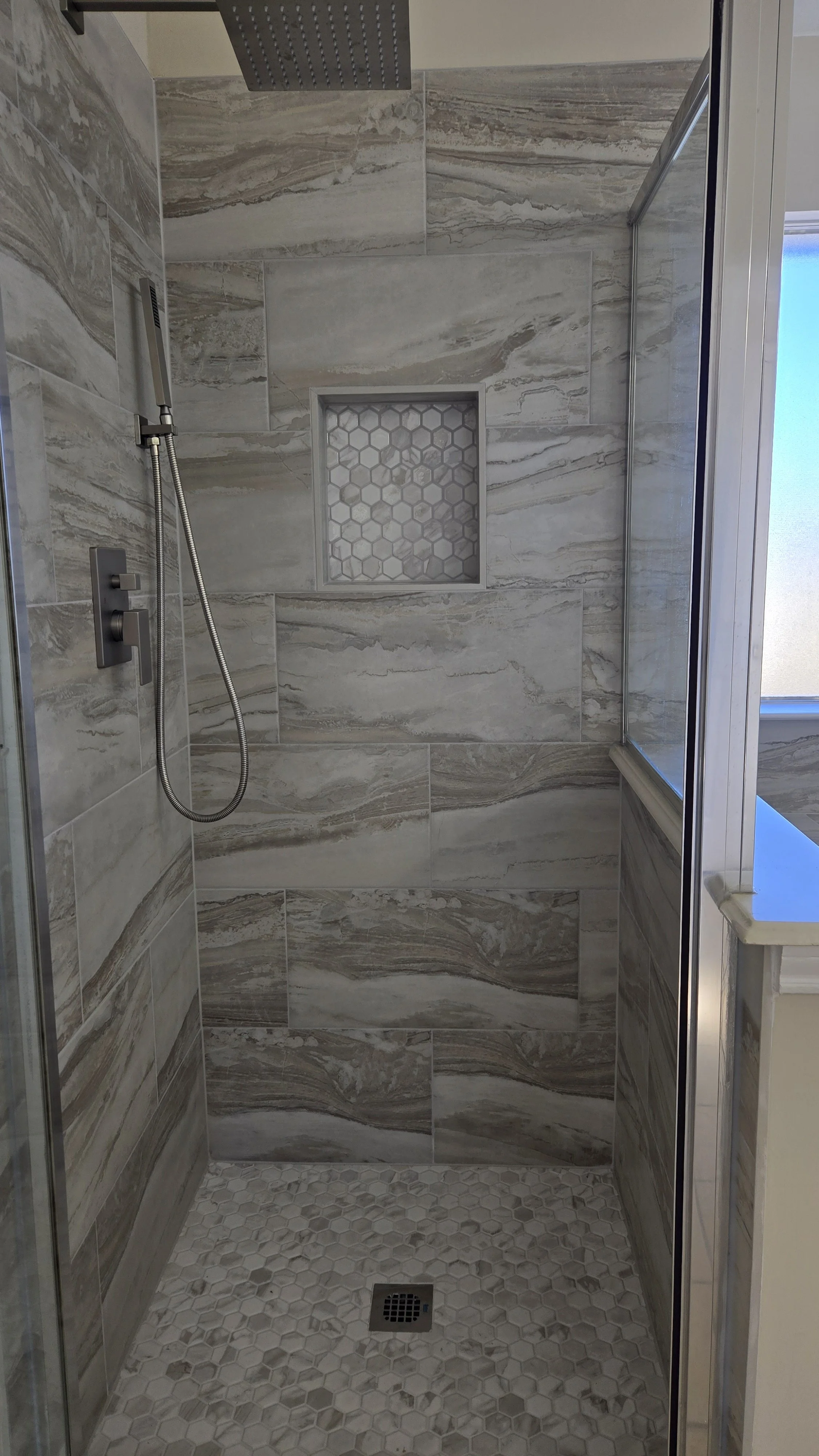 TurnKey Roofing & Restoration — A newly renovated / remodeled modern walk-in shower with large marble-look tiles, a built-in hexagonal tile niche, small hexagonal floor tiles, and a high shower head. Houston, Texas