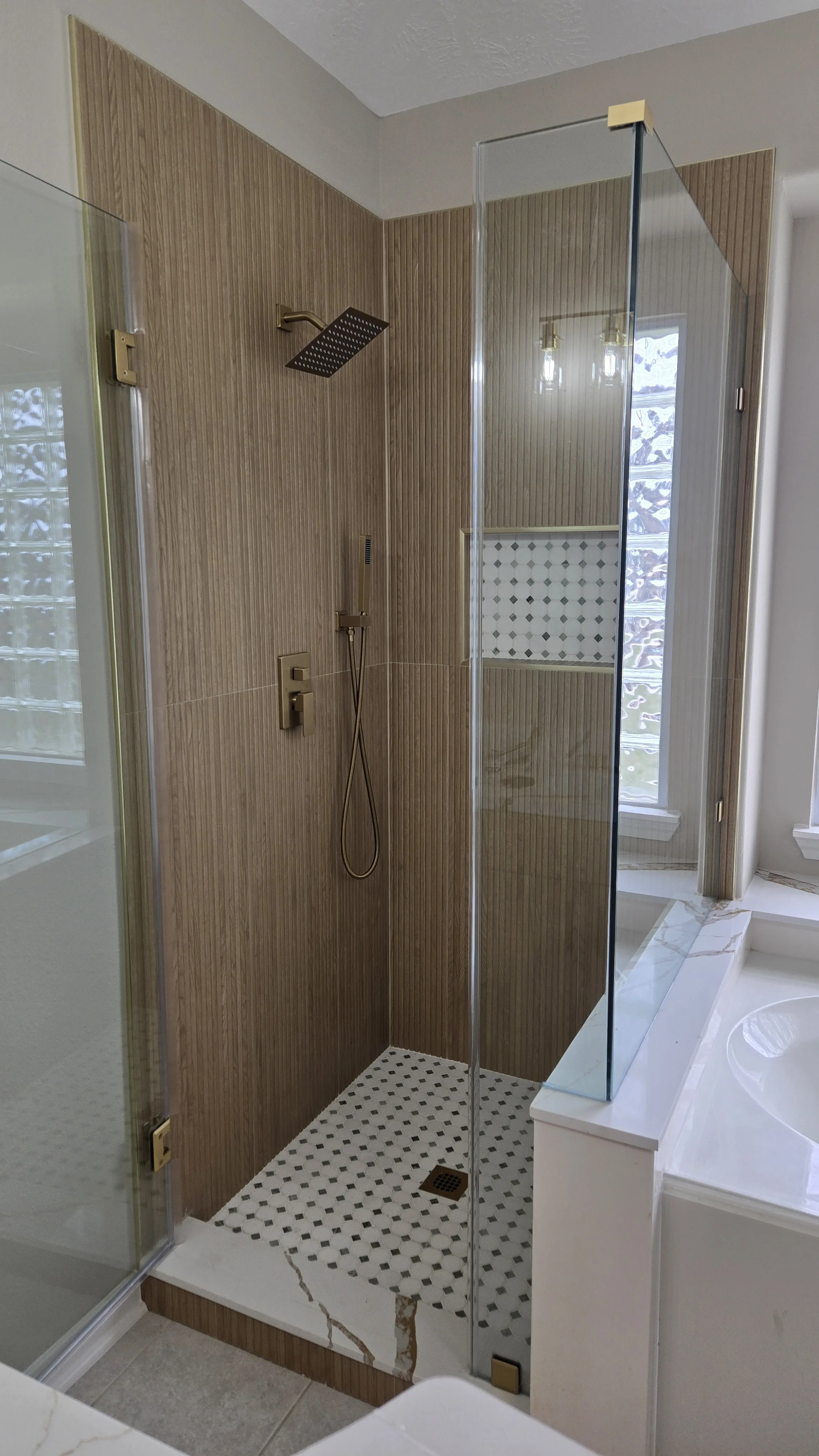 TurnKey Roofing & Restoration — A newly renovated / remodeled modern bathroom shower with a black square rainfall showerhead, a handheld shower with hose, wooden panel wall, a glass door, and a mosaic tile floor. Houston, Texas