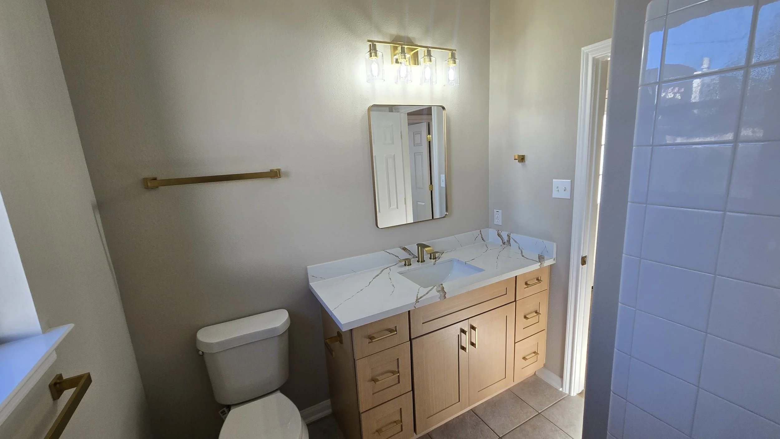 	
TurnKey Roofing & Restoration — A remodeled bathroom with a wooden vanity, a marble countertop, a rectangular mirror, gold fixtures, a white toilet, a gold towel rack, a wall-mounted light fixture, and a tiled shower area with a glass block wall.