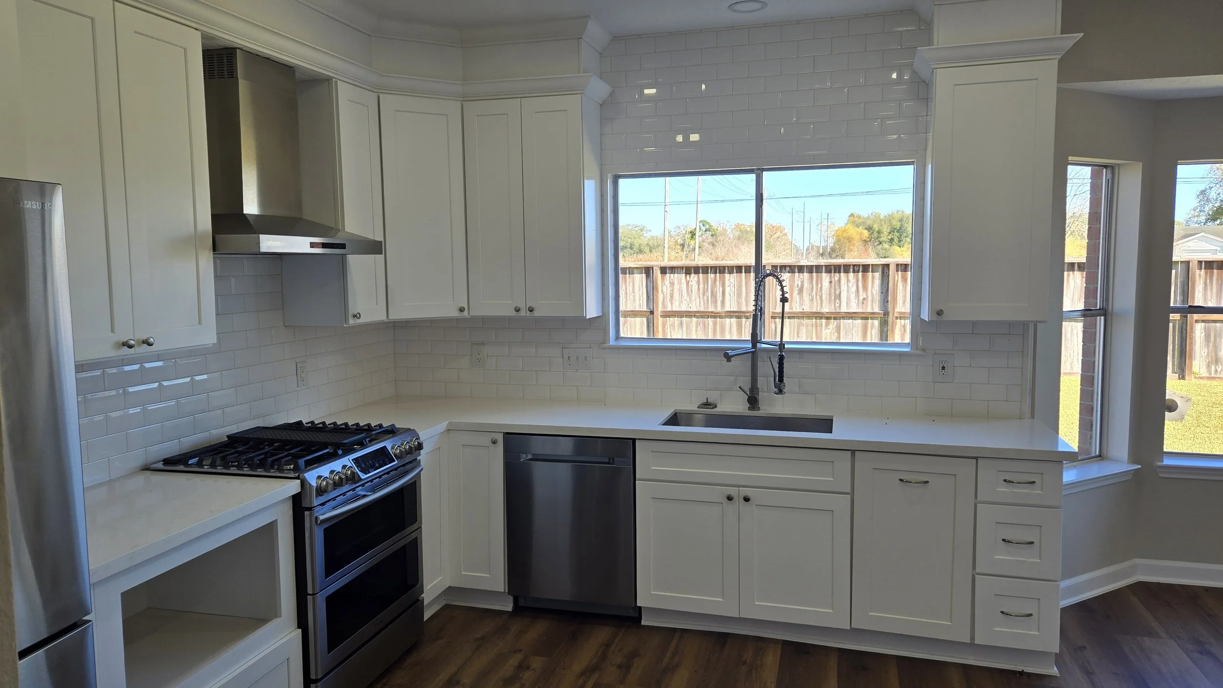 TurnKey Roofing & Restoration — A newly renovated / remodeled modern kitchen with white cabinets, a stainless steel refrigerator, a black stove, white subway tile backsplash, and a large window over the sink revealing the yard outside. Houston, Texas
