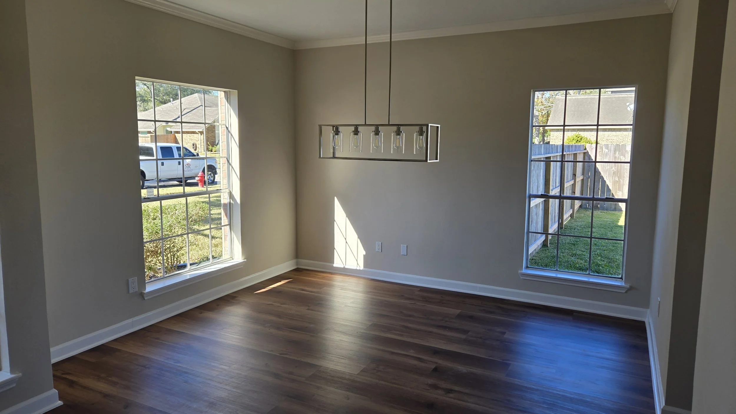 TurnKey Roofing & Restoration — A newly renovated / remodeled dining room with wooden floors, beige walls, two large windows, and a modern rectangular chandelier with exposed bulbs hanging in the center. Houston, Texas